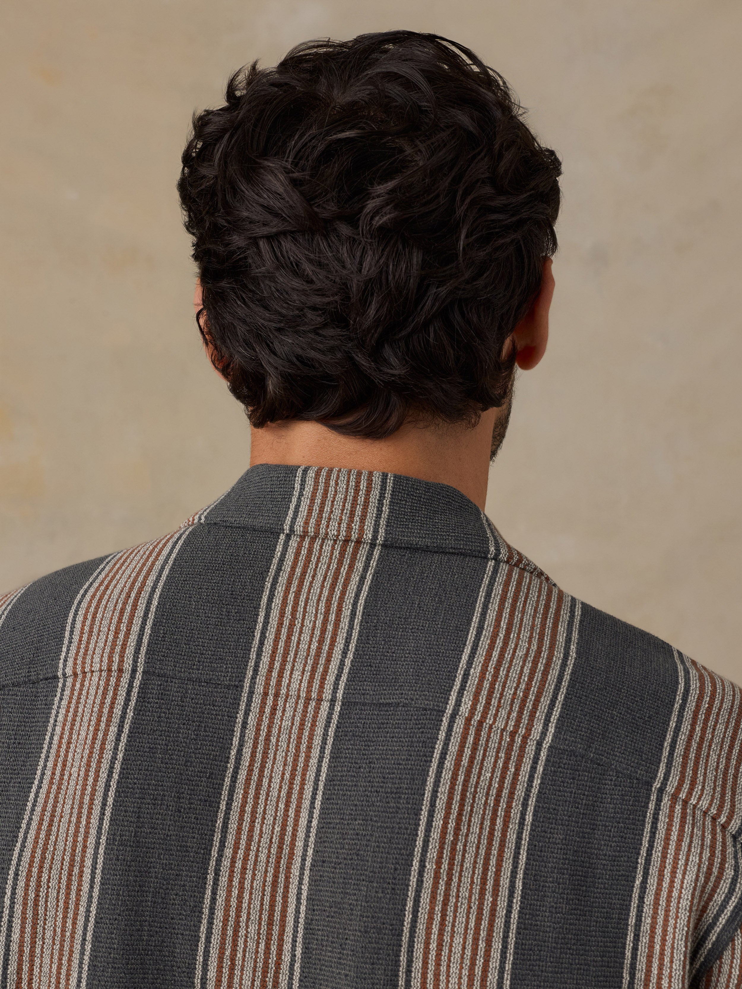 Back view of a man with dark curly hair wearing a gray striped blazer with vertical stripes in beige, brown, and white, set against a neutral beige background.