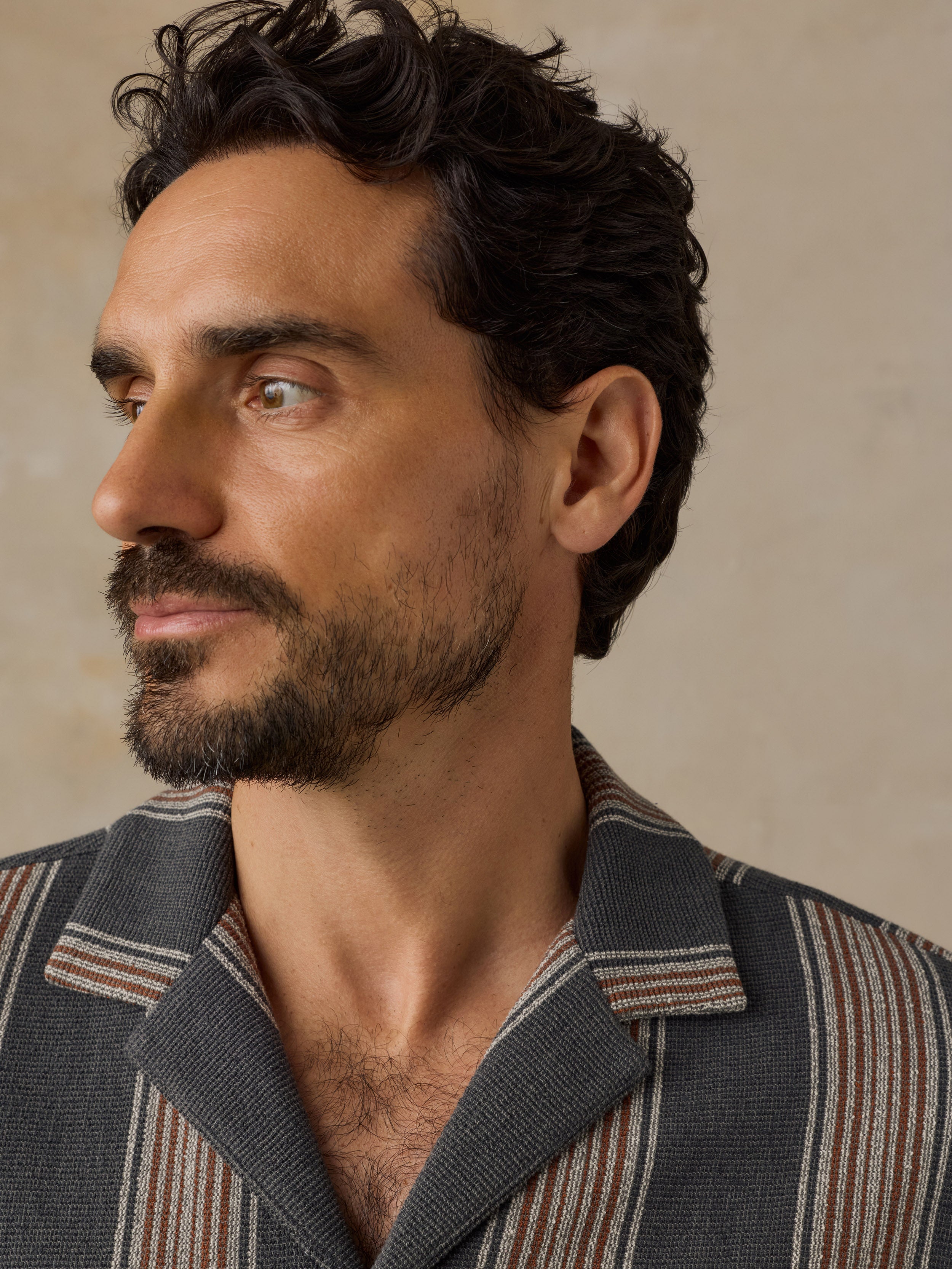 A close-up portrait of a man wearing a textured gray striped button-down shirt with brown and beige vertical stripes, set against a neutral beige background.