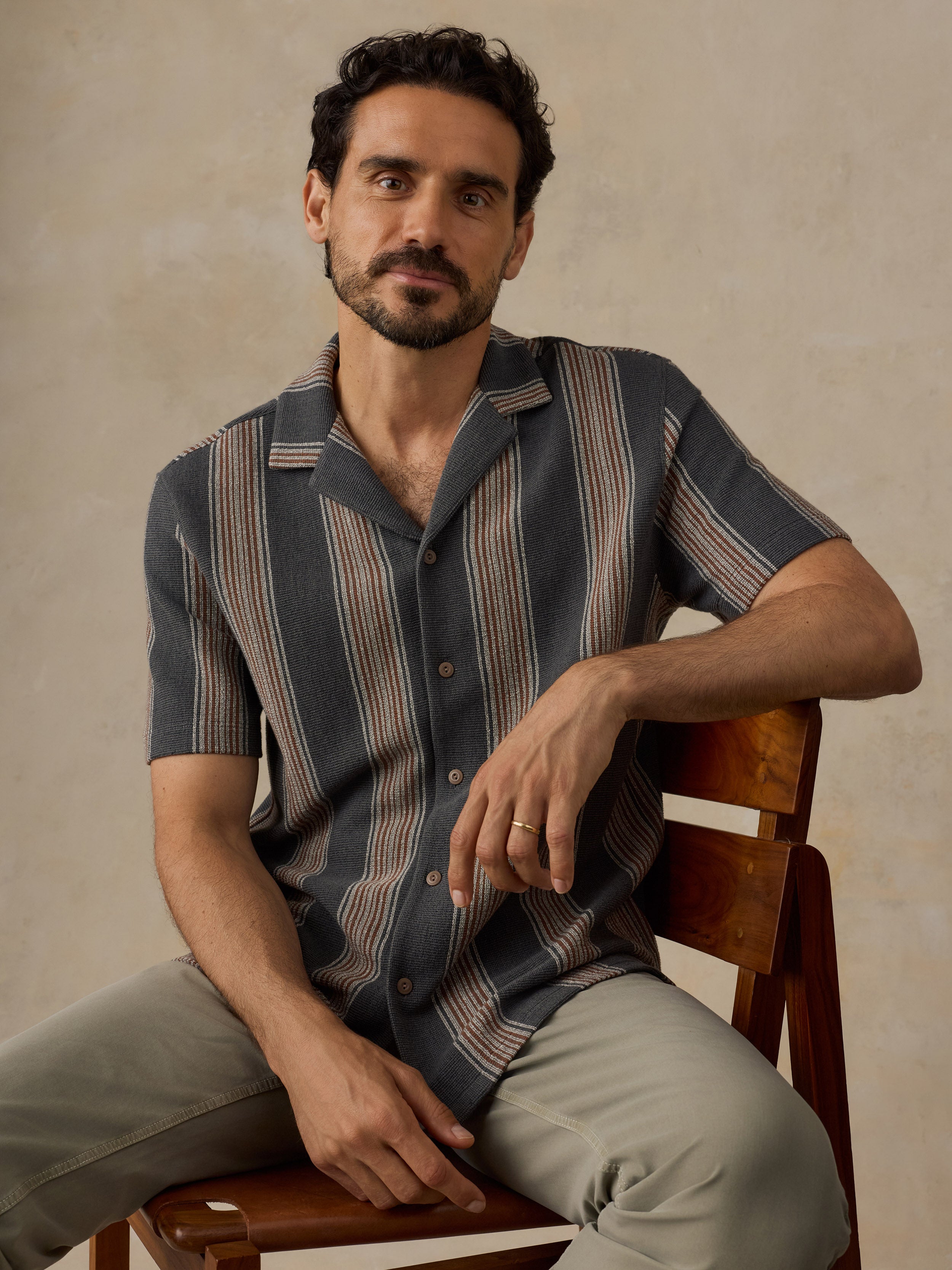 A man wearing a short-sleeve button-up shirt with vertical stripes in slate blue, beige, and brown, paired with light gray trousers, sitting on a wooden chair against a textured beige wall.
