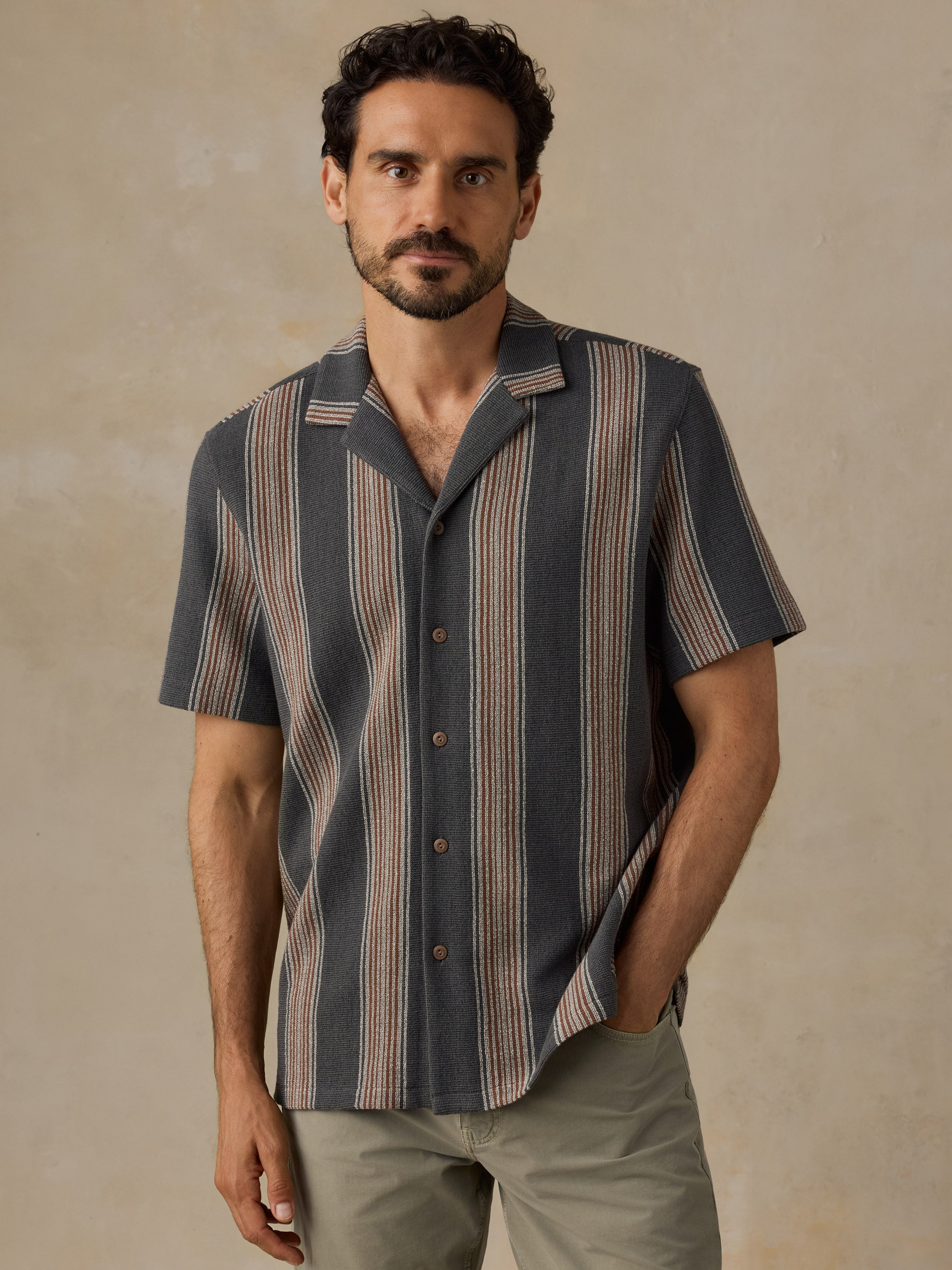 A man wearing a short-sleeve button-up shirt with vertical stripes in slate blue, beige, and brown, paired with khaki pants, standing against a textured beige wall.