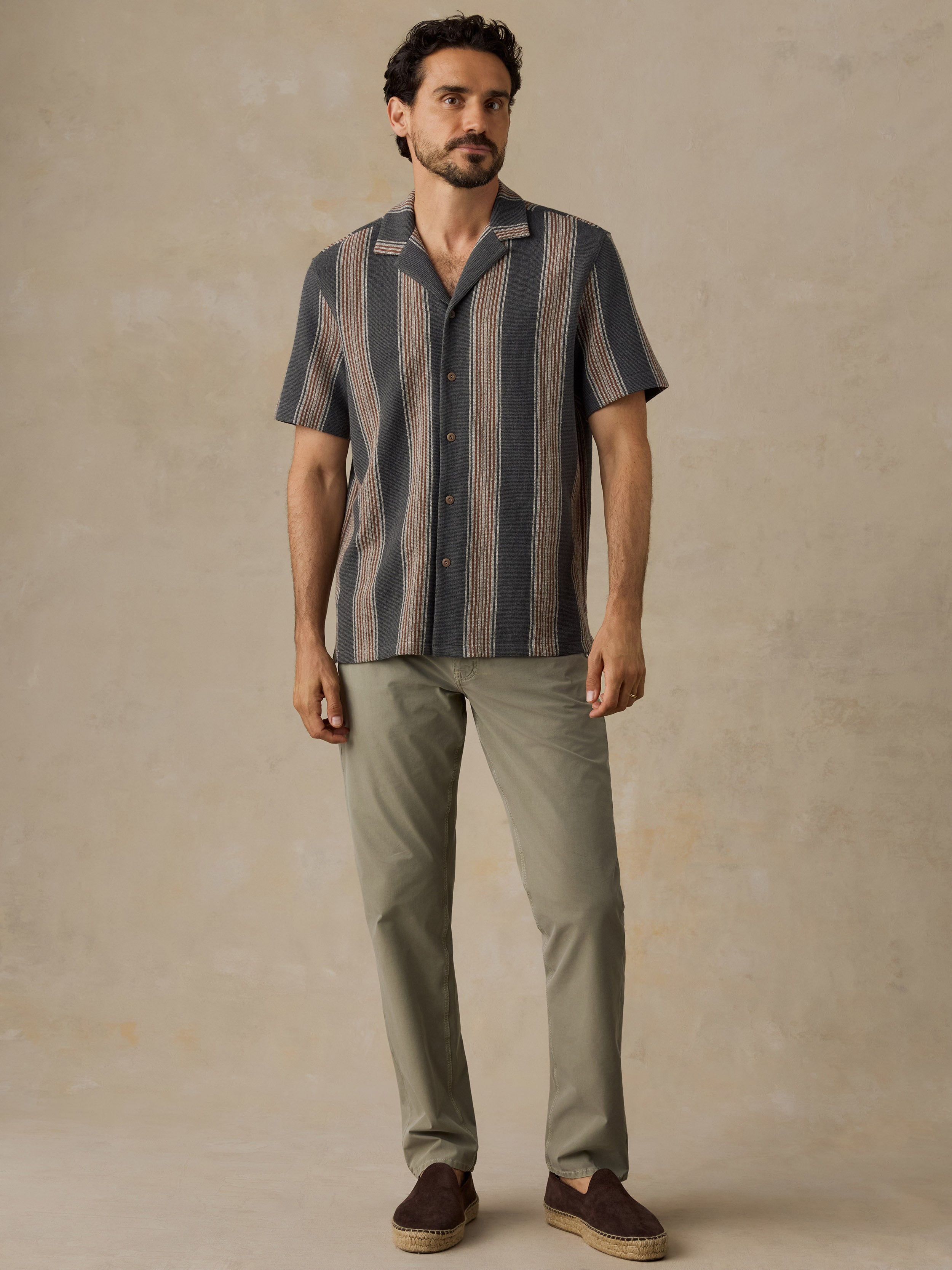A man modeling a short-sleeve button-up shirt with vertical stripes in navy, gray, and beige, paired with olive green trousers and brown slip-on shoes, against a textured beige studio backdrop.