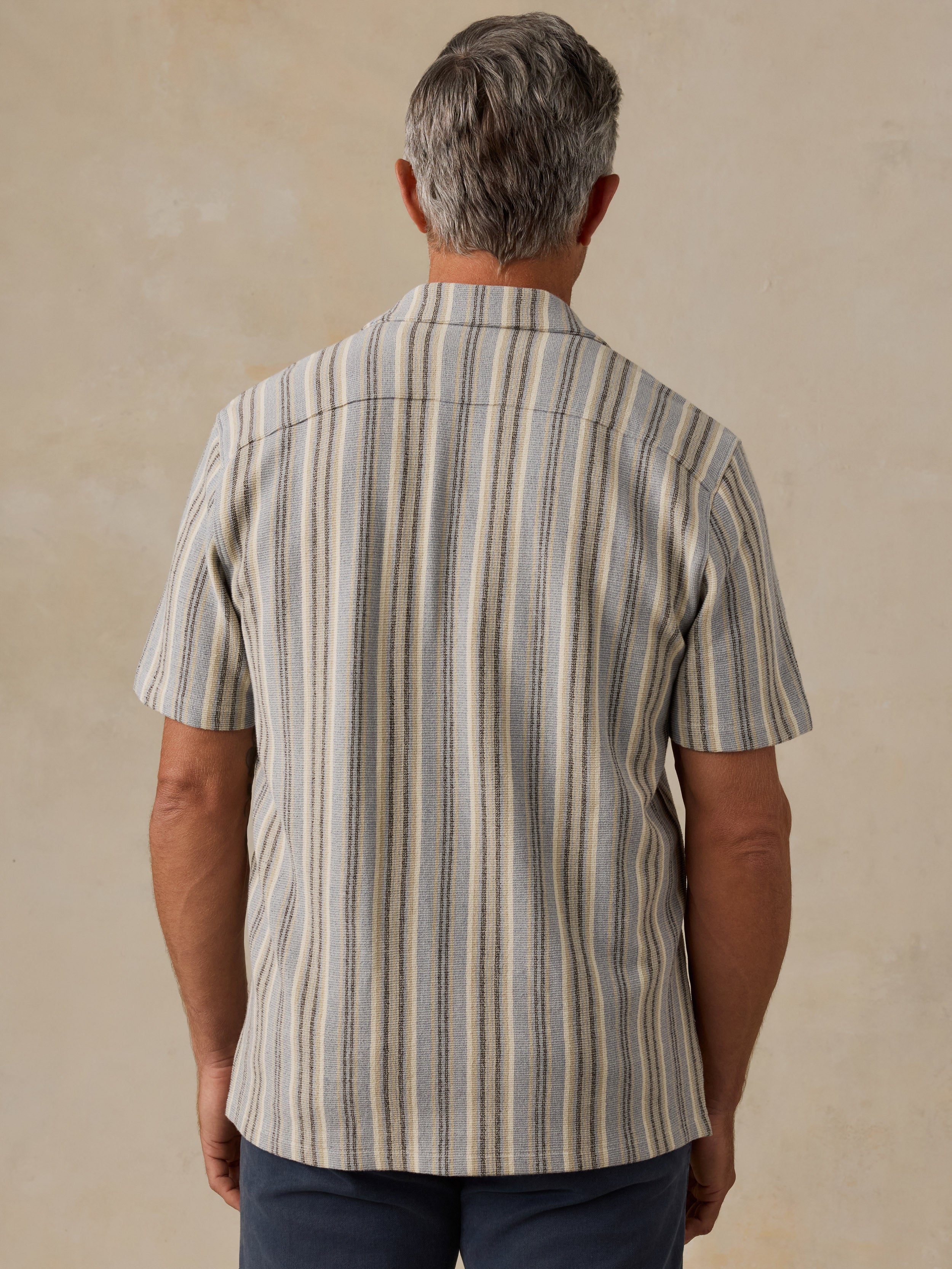A man viewed from behind wearing a short-sleeve button-up shirt with vertical stripes in beige, gray, and brown tones, paired with dark blue pants, set against a textured beige background.
