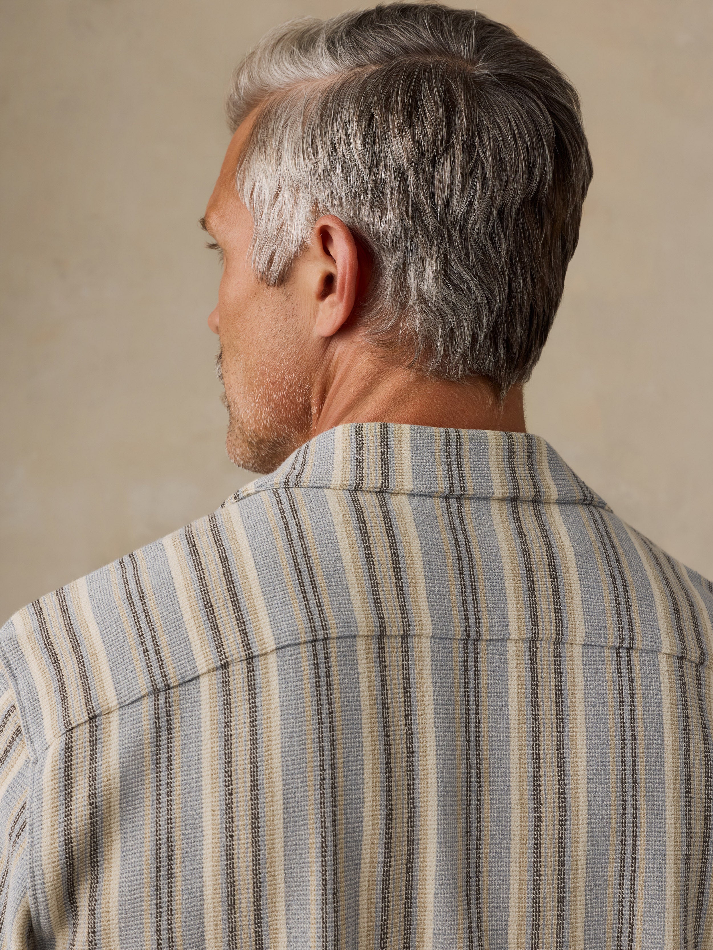A close-up back view of a man with gray hair wearing a textured striped button-down shirt in beige, gray, and navy blue.