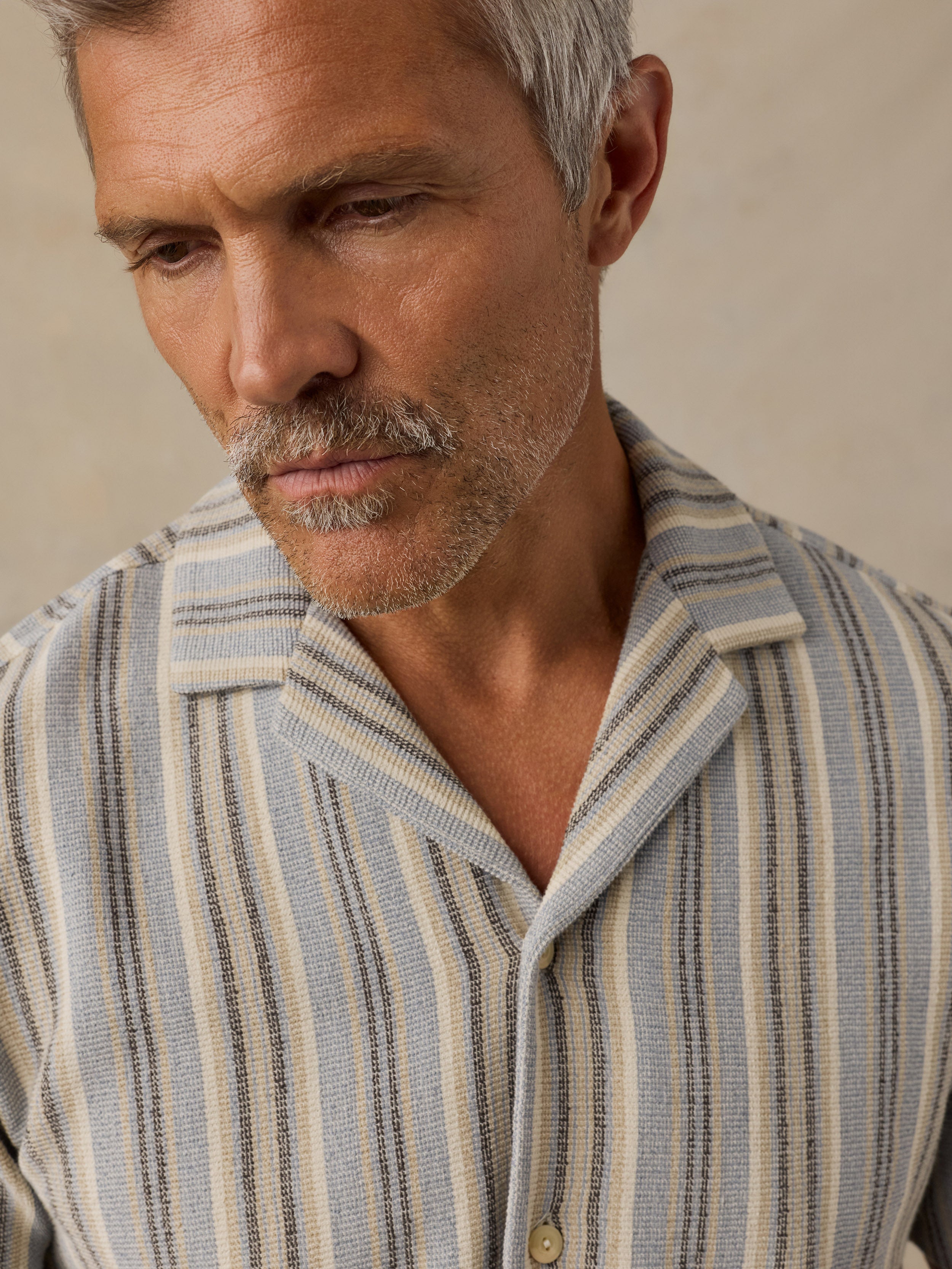 A close-up of a man wearing a textured, vertically striped button-down shirt in beige, light blue, and dark gray.