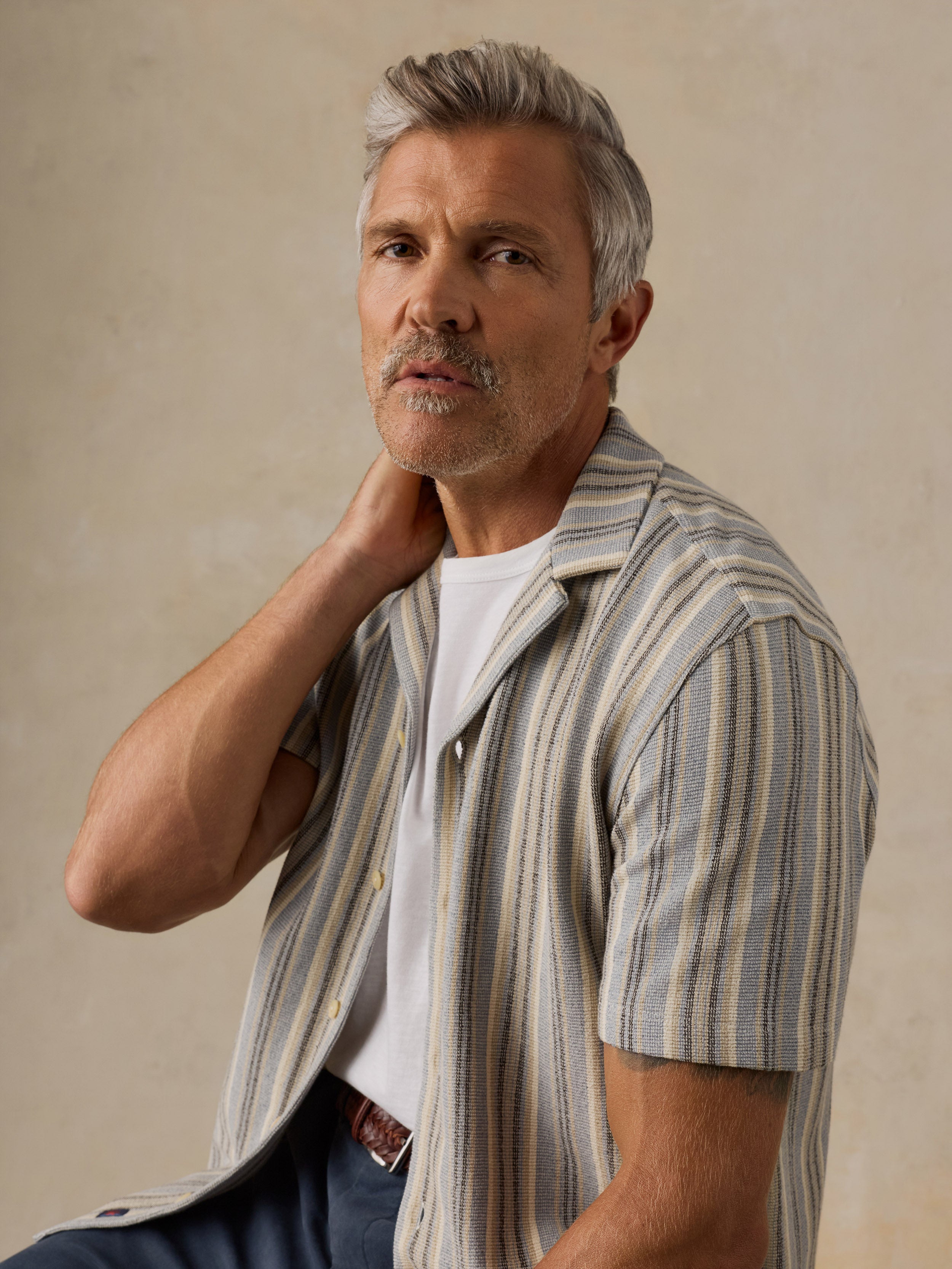 A man with gray hair and a mustache wearing a short-sleeve button-up shirt with vertical beige, gray, and white stripes over a white t-shirt, posed against a neutral textured beige background.
