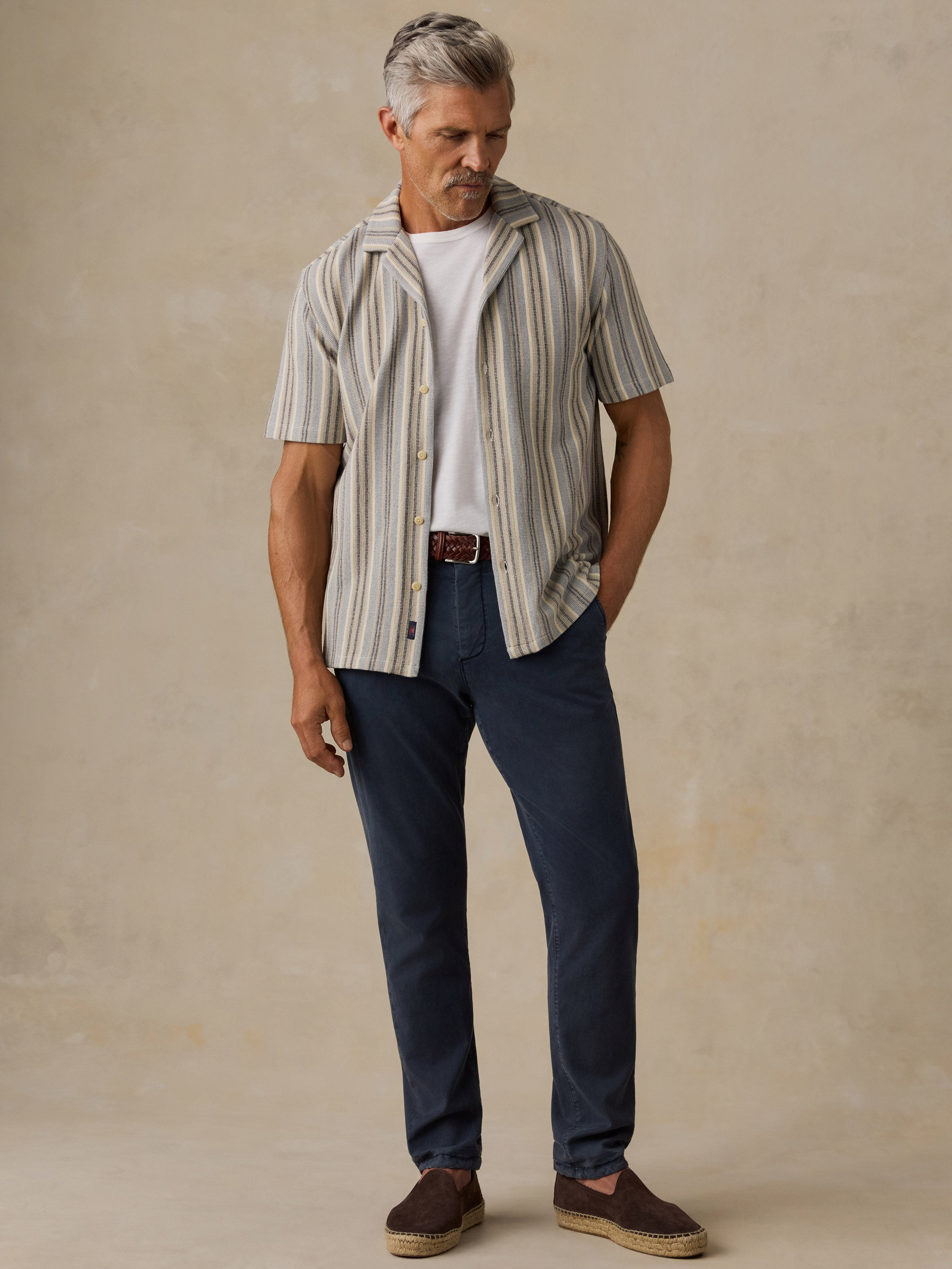 A man wearing a beige and gray vertically striped short-sleeve button-up shirt over a white t-shirt, paired with navy blue jeans and brown slip-on shoes, standing against a textured beige studio backdrop.