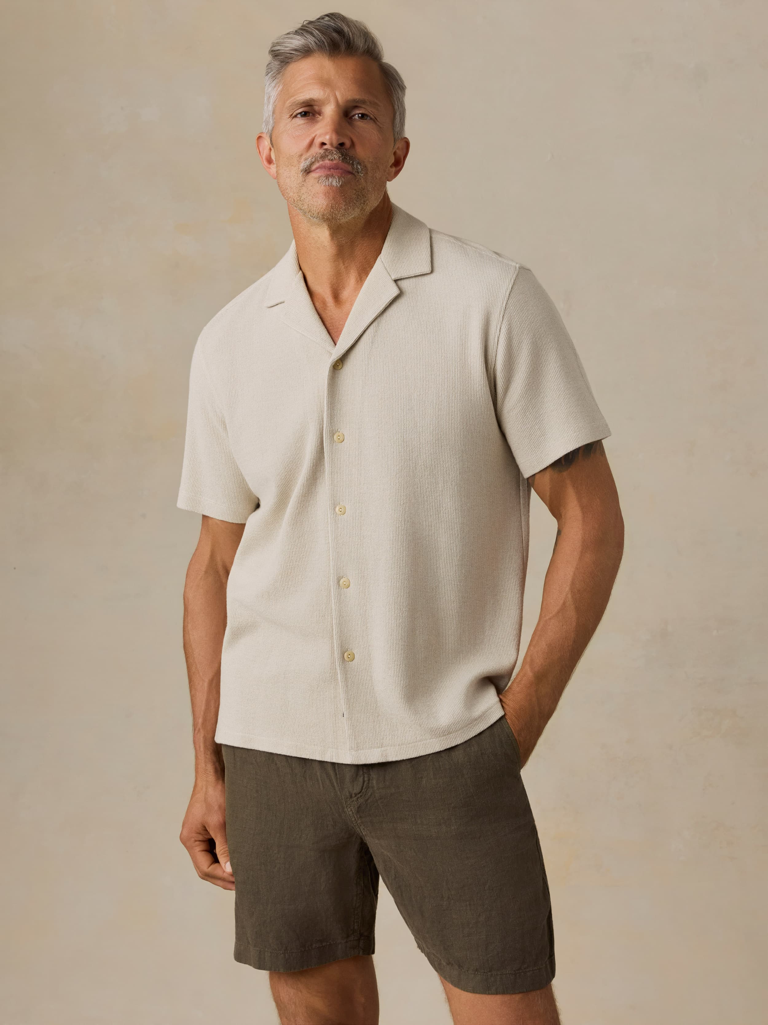 Man wearing a beige short-sleeve shirt and dark shorts against a beige background