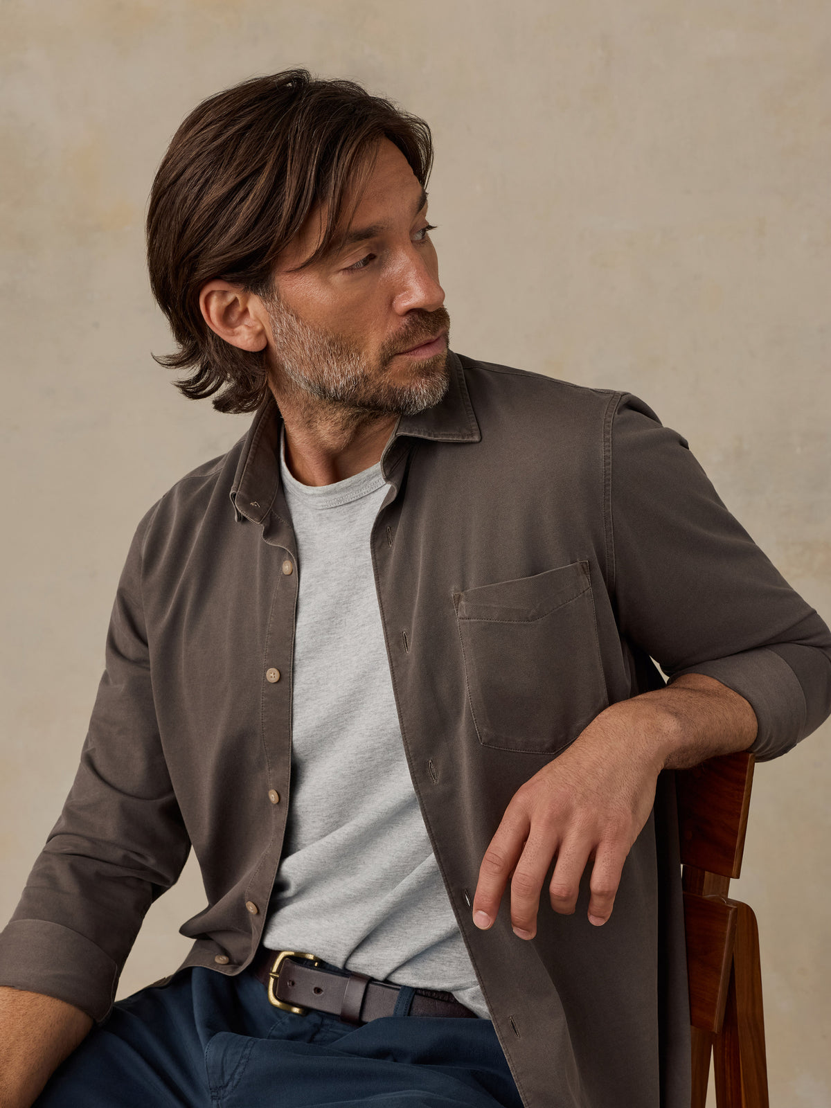 Man sitting on a wooden chair wearing a brown button down shirt and navy chino pants against a beige background, image 2 of 5