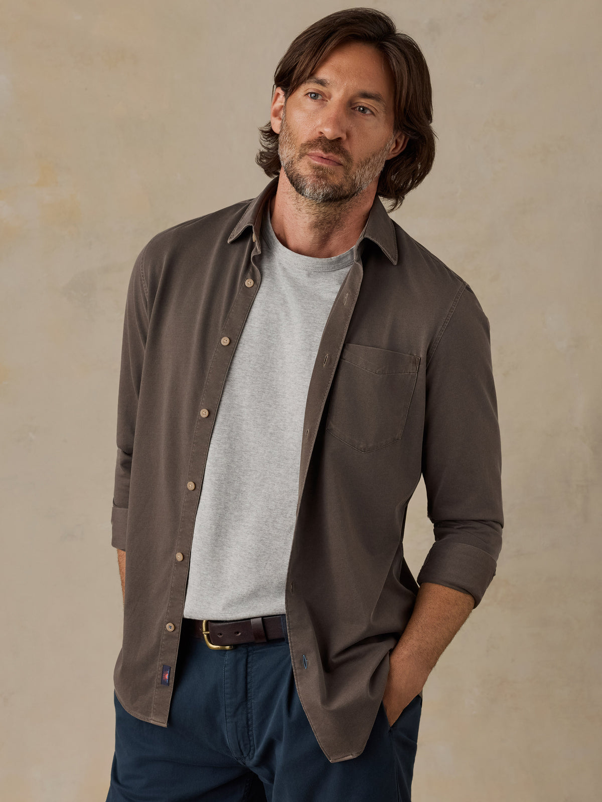 Man wearing a brown button down shirt and navy chino pants against a beige background, image 1 of 5
