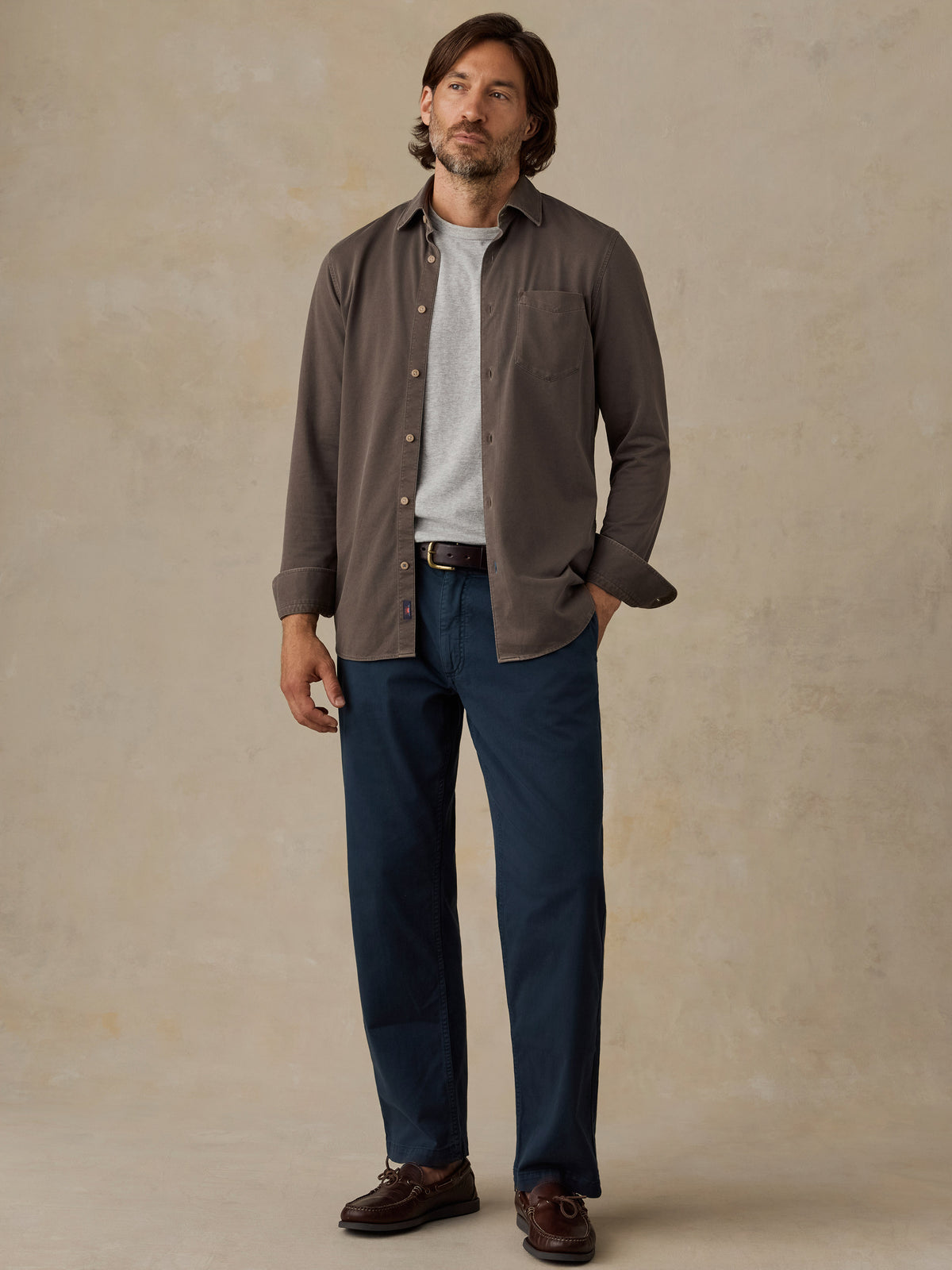Man wearing a brown button down shirt and navy chino pants against a beige background, image 5 of 5