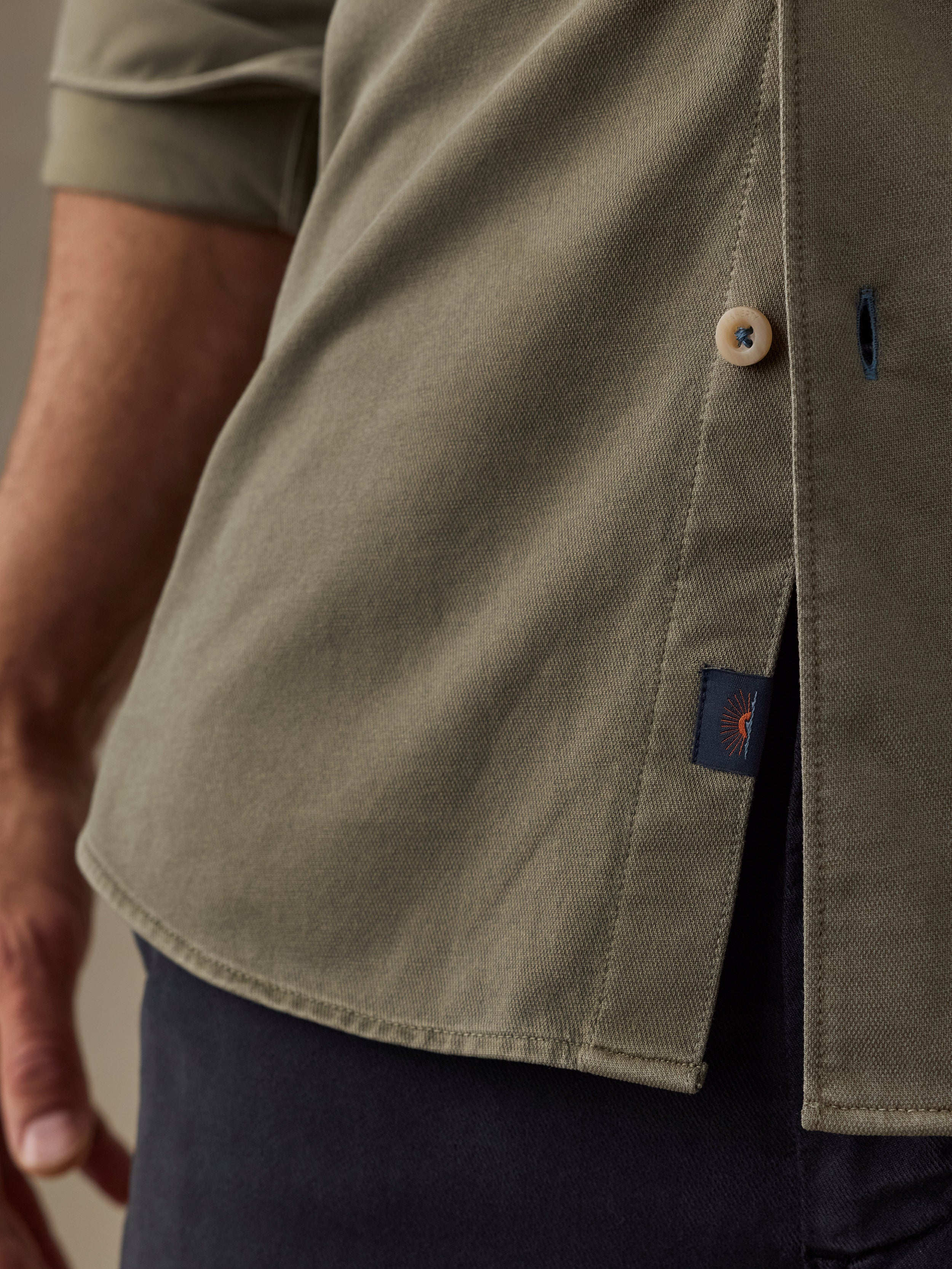 Close-up of a man wearing an olive green short-sleeve button-up shirt with a side slit and a small navy Faherty brand tab near the hem.