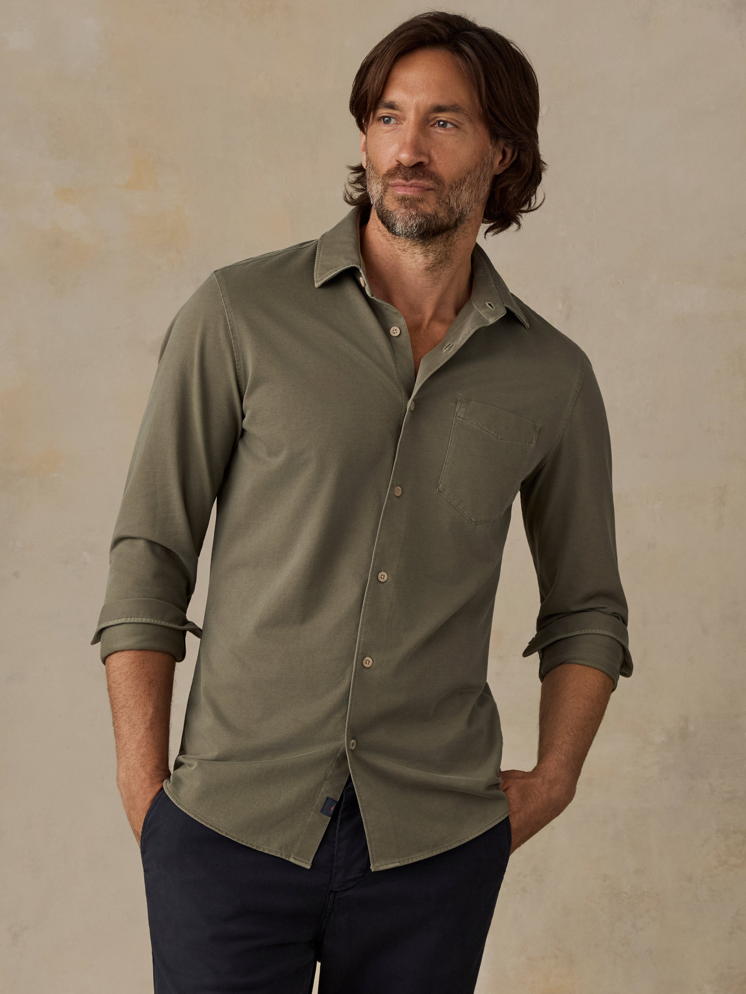 A man wearing a muted olive green long-sleeve button-down shirt with rolled-up sleeves and dark navy pants, standing against a textured beige studio backdrop.