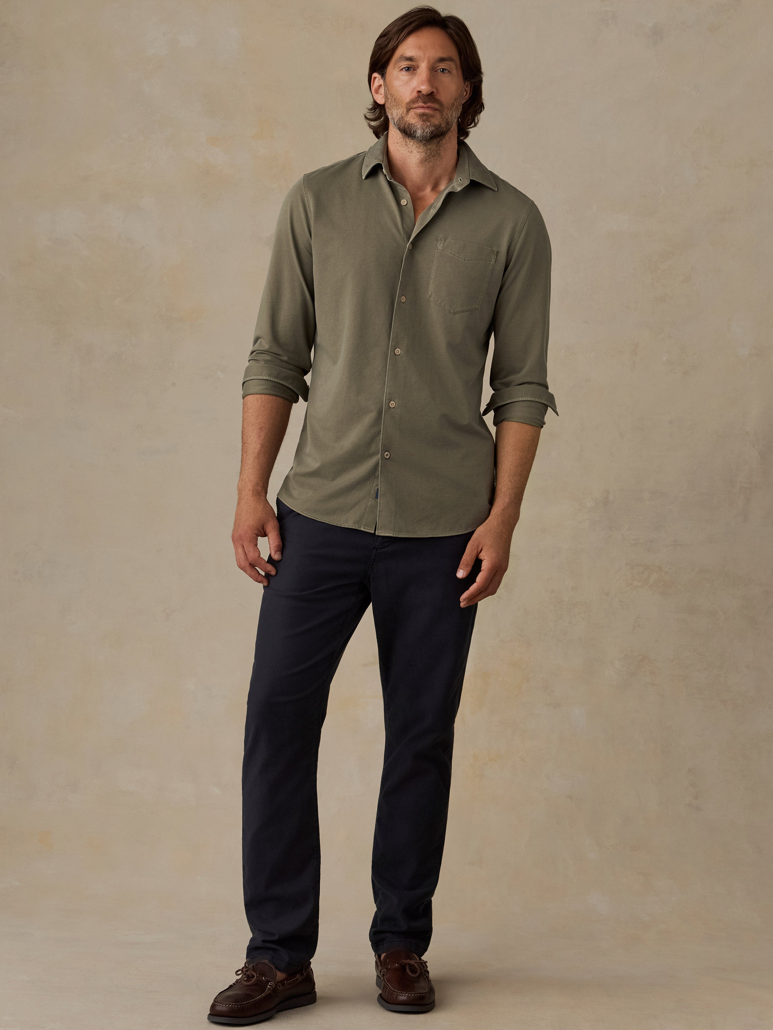A man wearing an olive green long-sleeve button-down shirt with dark navy pants and brown shoes, standing against a textured beige studio backdrop.