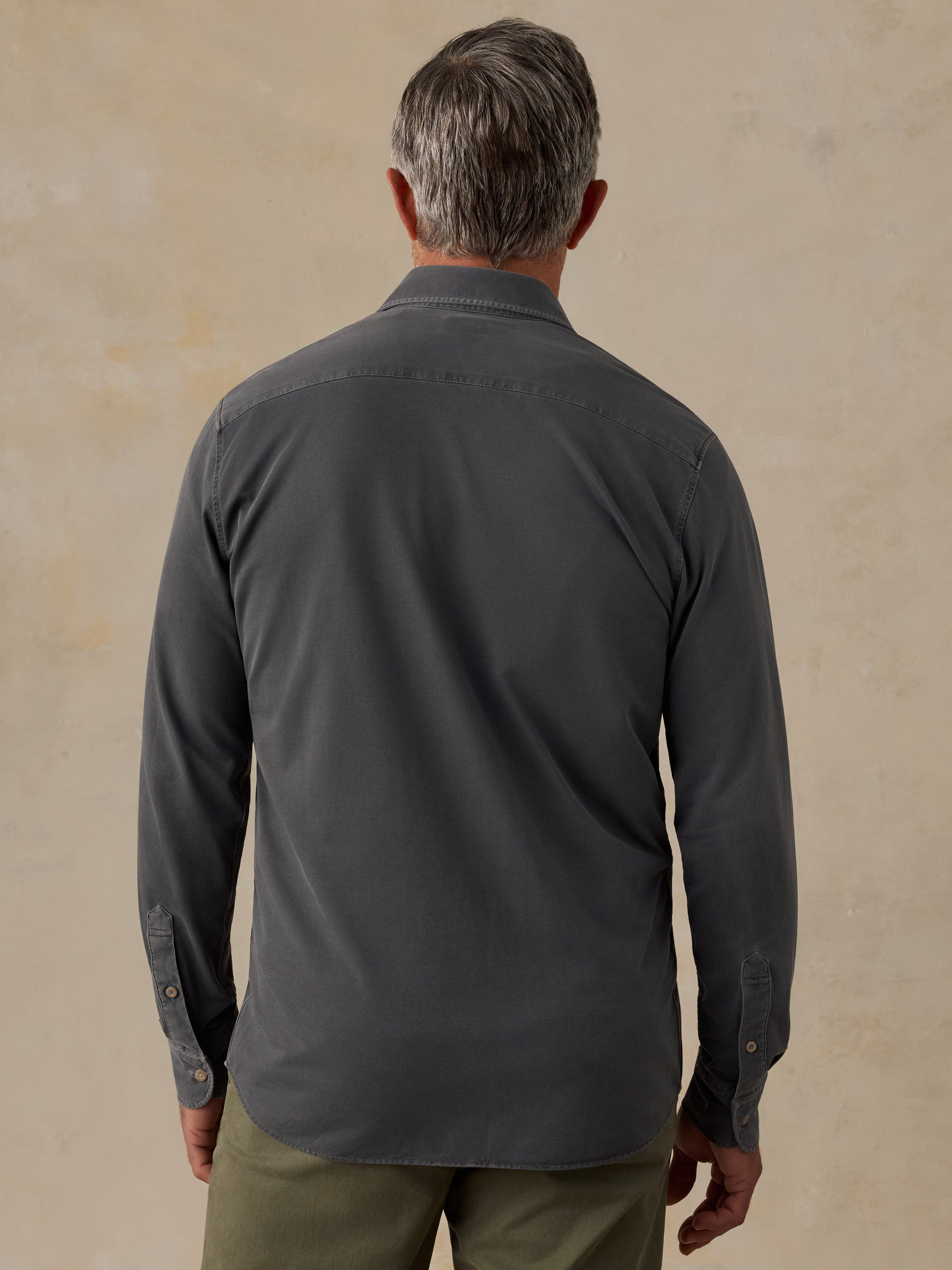 A man viewed from behind wearing a dark charcoal gray long-sleeve button-down shirt and olive green pants, standing against a textured beige background.