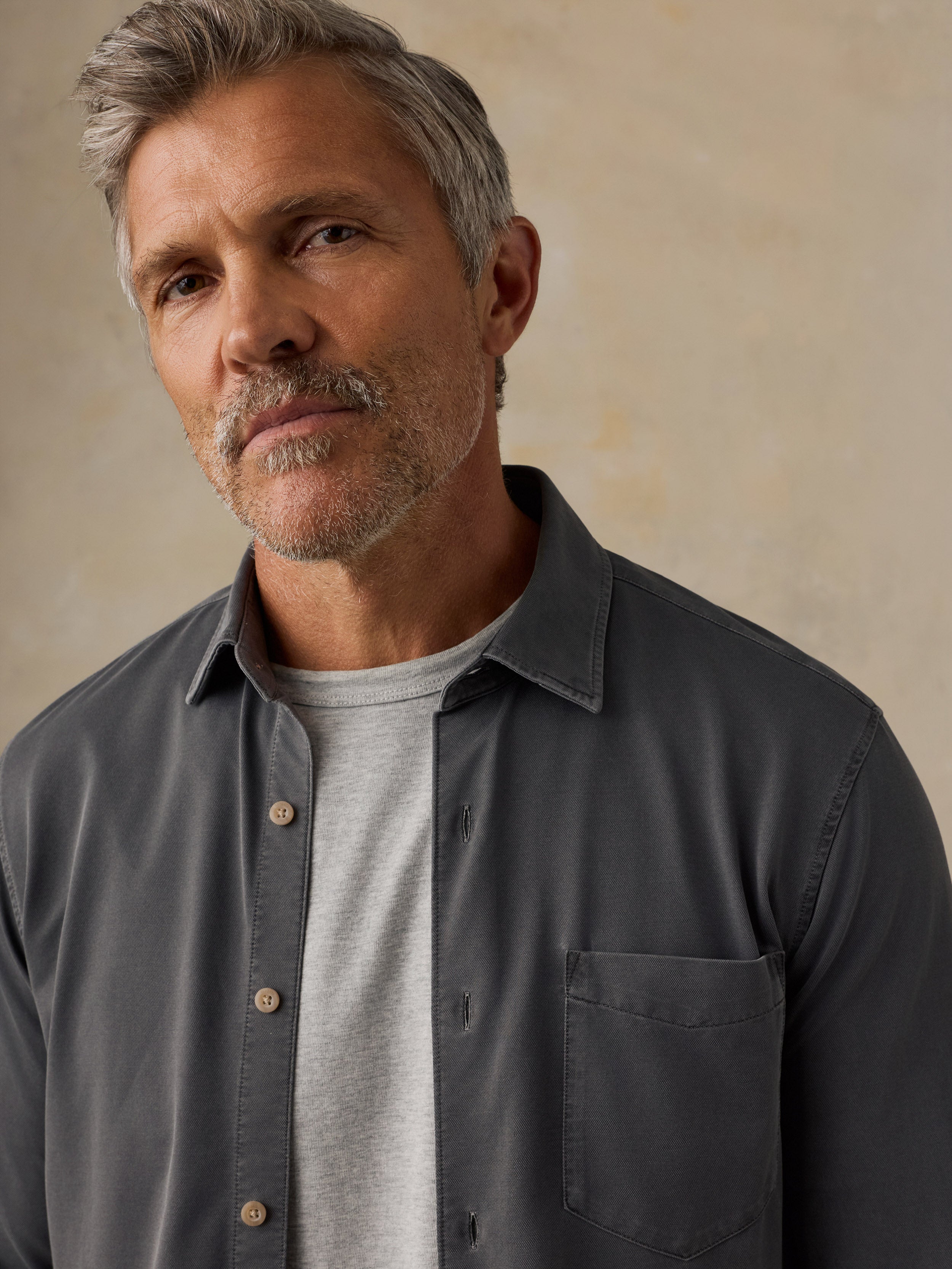 A close-up of a man wearing a dark charcoal gray button-down shirt over a light gray t-shirt, set against a warm beige textured background.