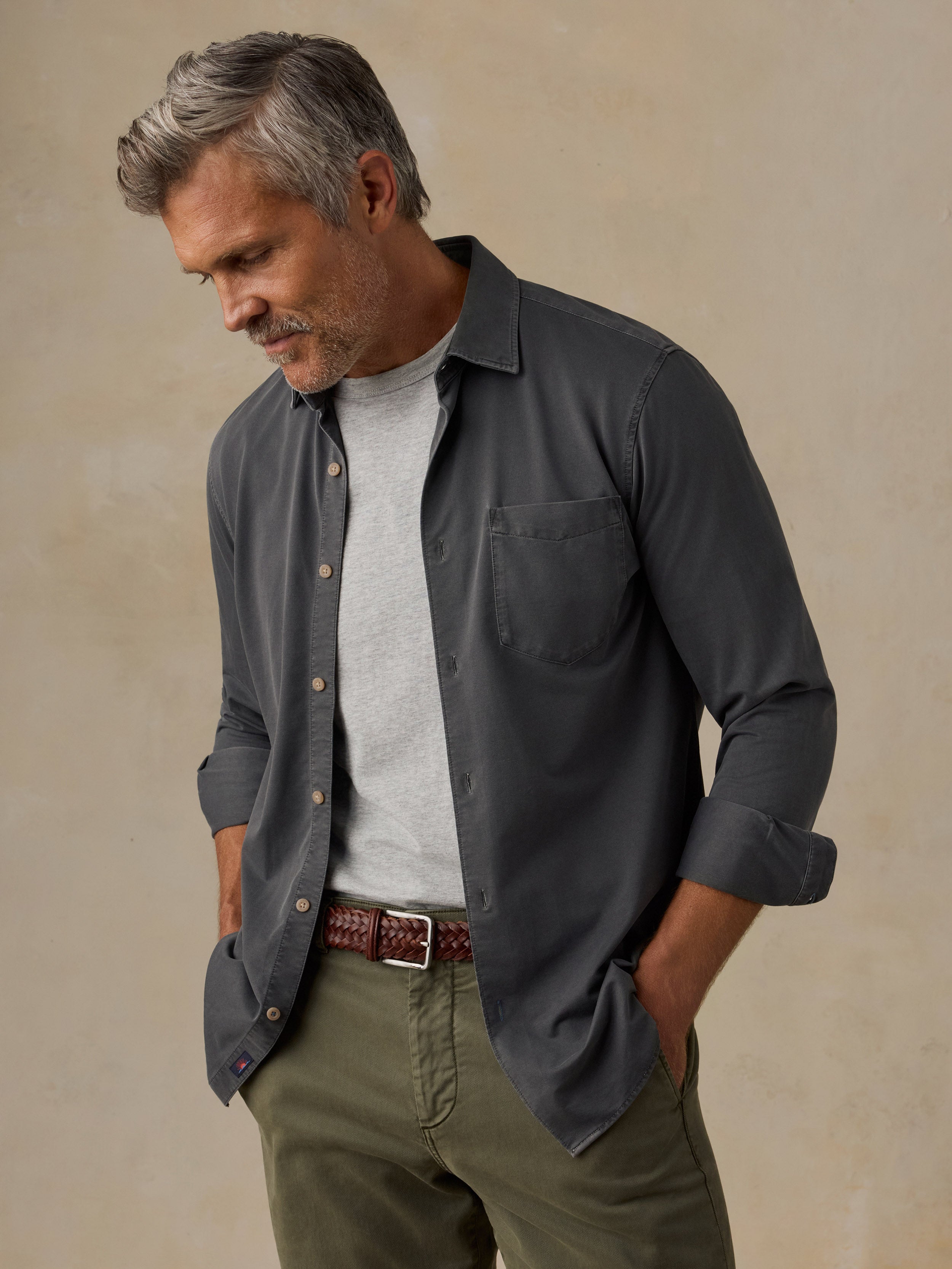 A man wearing a dark charcoal gray long-sleeve button-down shirt over a light gray t-shirt, paired with olive green pants and a brown belt, standing against a textured beige background.