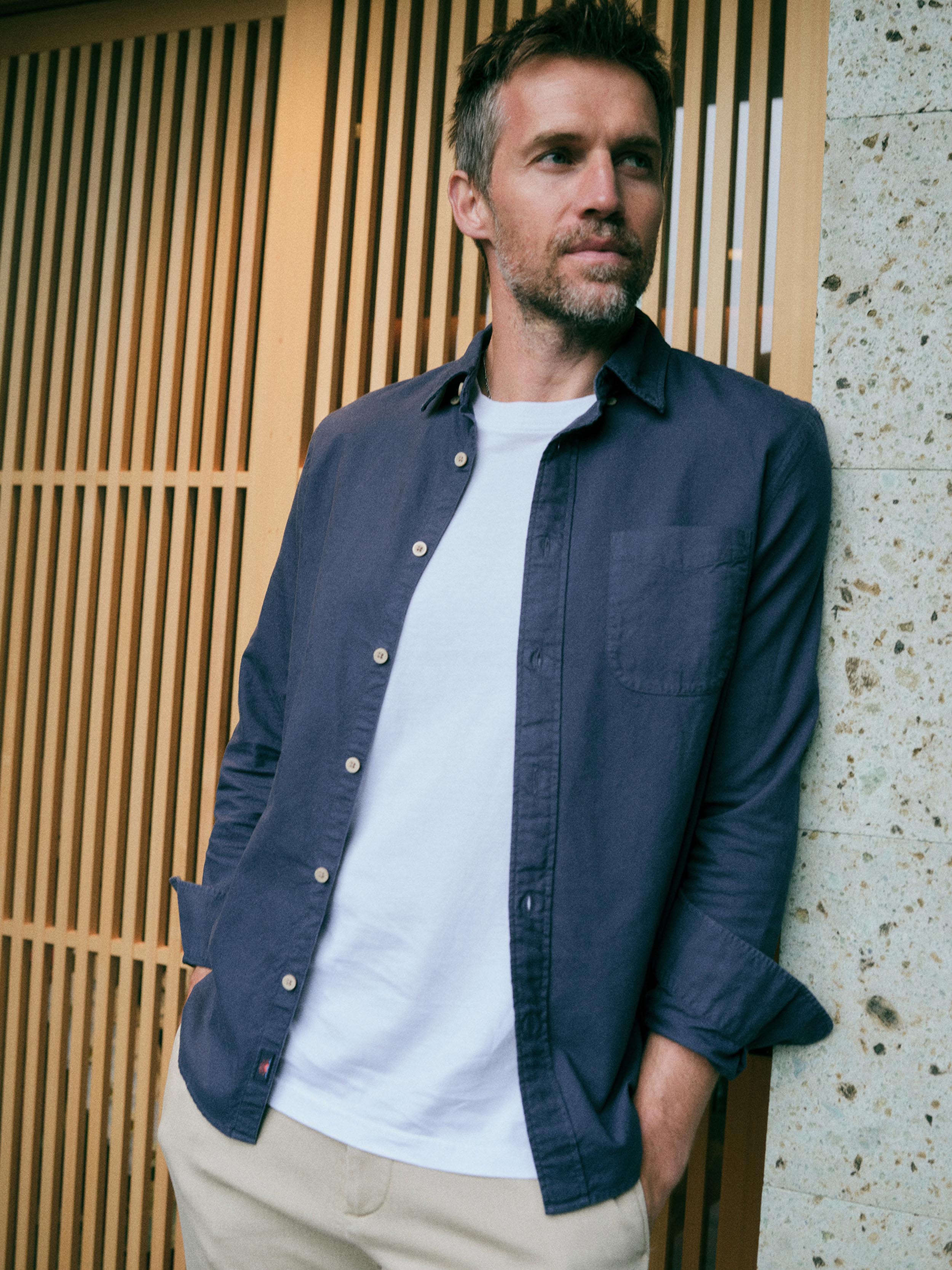 A man wearing a navy blue long-sleeve button-down shirt over a white t-shirt and beige pants, leaning against a modern wooden slat wall and concrete pillar.