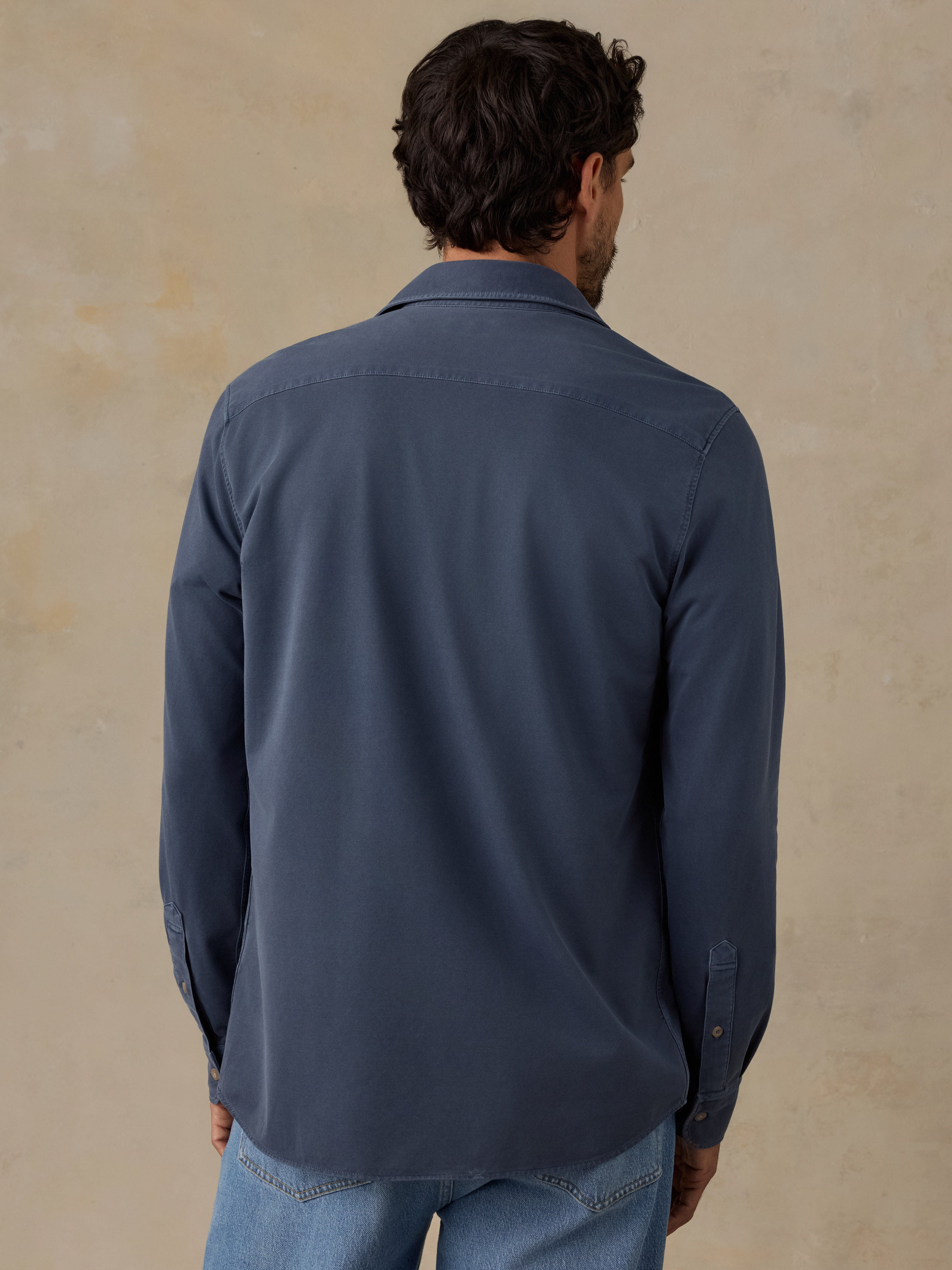 A man viewed from behind wearing a slate blue long-sleeve button-down shirt and light blue jeans, standing against a textured beige wall.