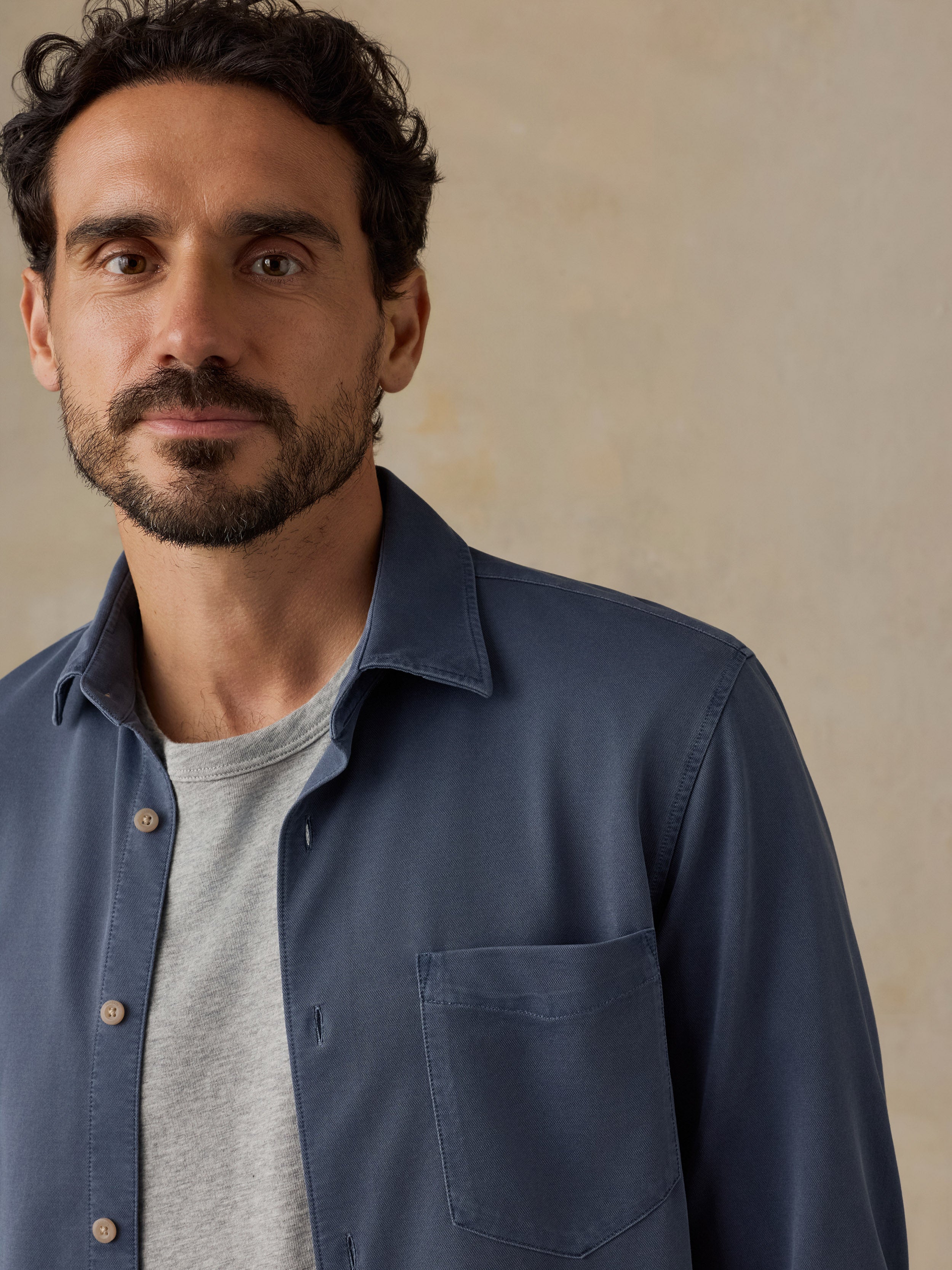 A close-up of a man wearing a slate blue long-sleeve button-down shirt over a light gray crew-neck t-shirt, set against a neutral textured beige background.