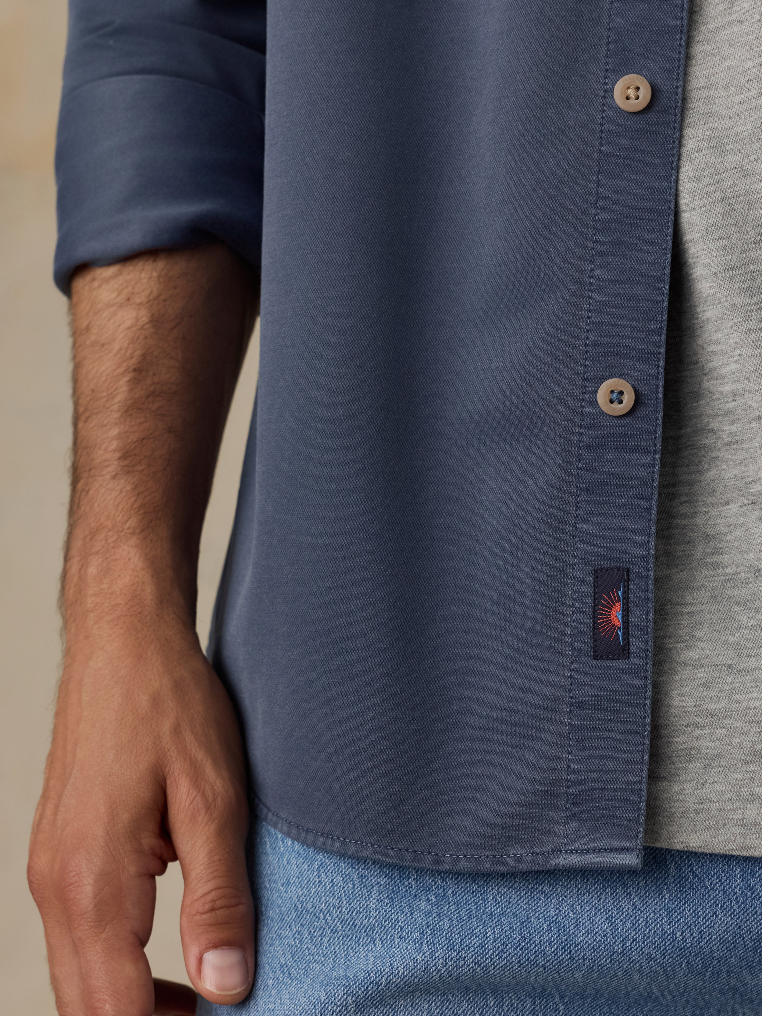 Close-up of a man wearing a blue long-sleeve button-up shirt over a heather gray T-shirt, with a small embroidered logo patch near the hem.