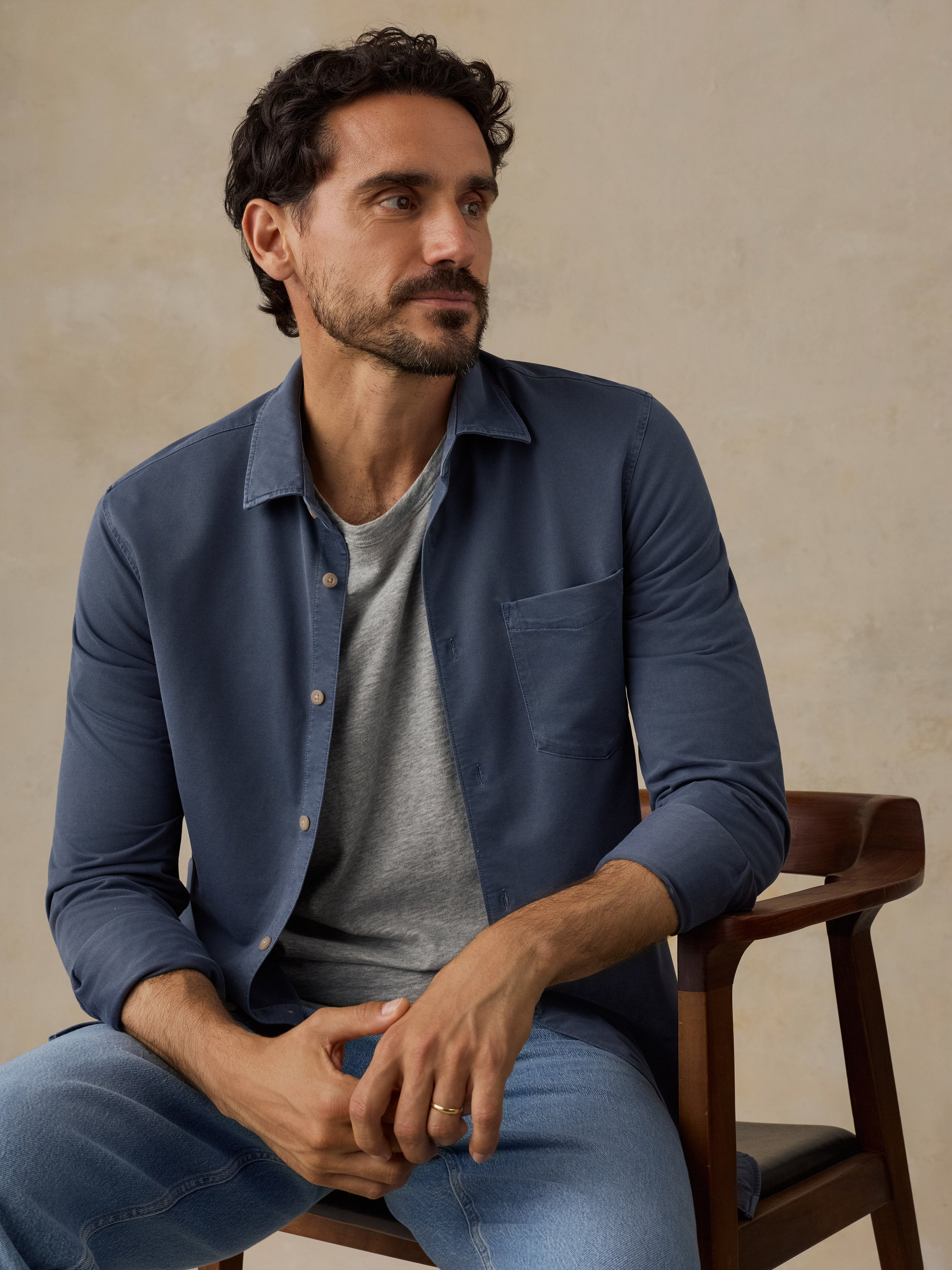 A man wearing a slate blue long-sleeve button-up shirt over a gray t-shirt and blue jeans, seated on a wooden chair against a textured beige wall.