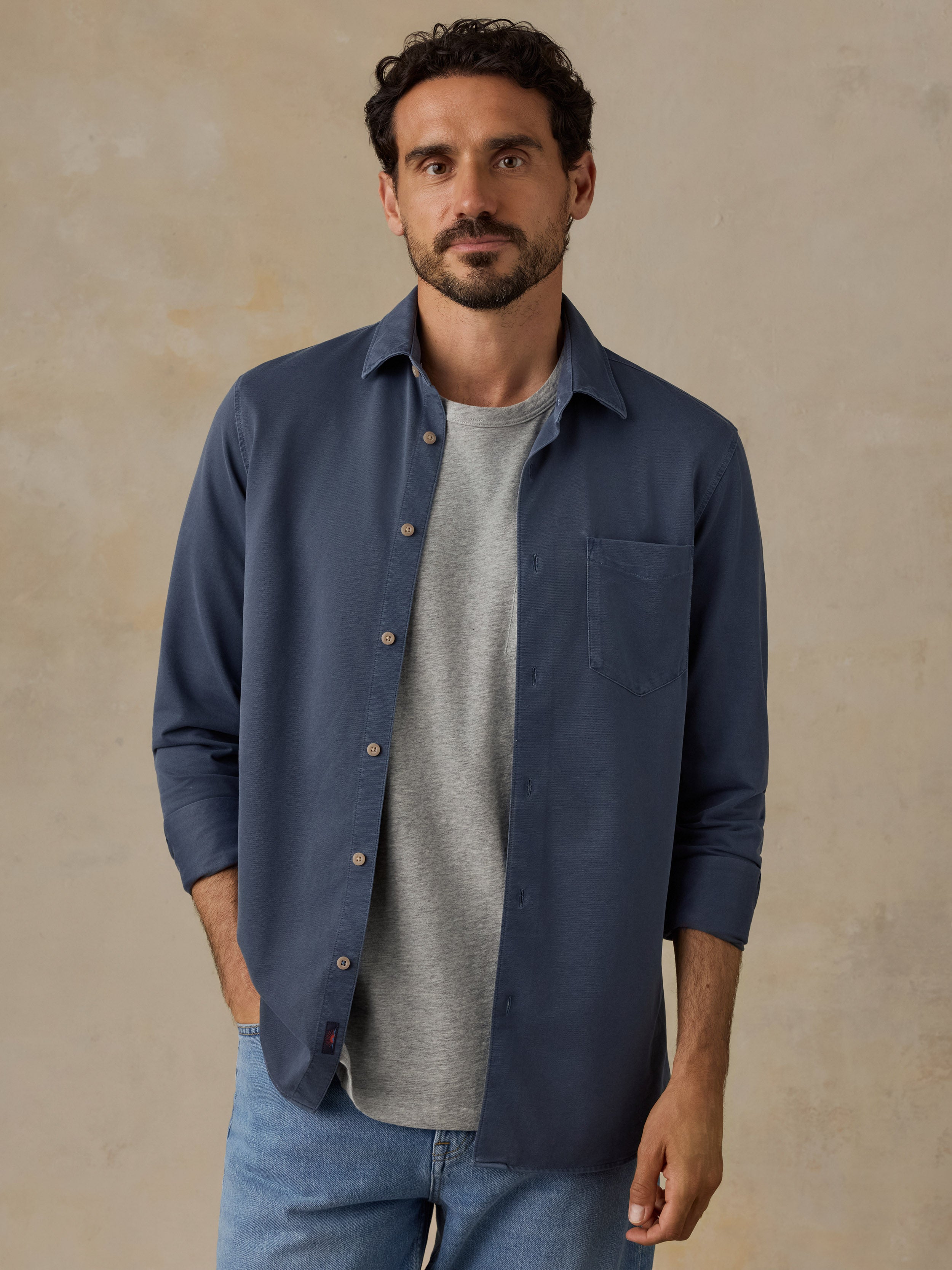 A man wearing a navy blue long-sleeve button-down shirt over a light gray t-shirt and blue jeans, standing against a textured beige studio background.