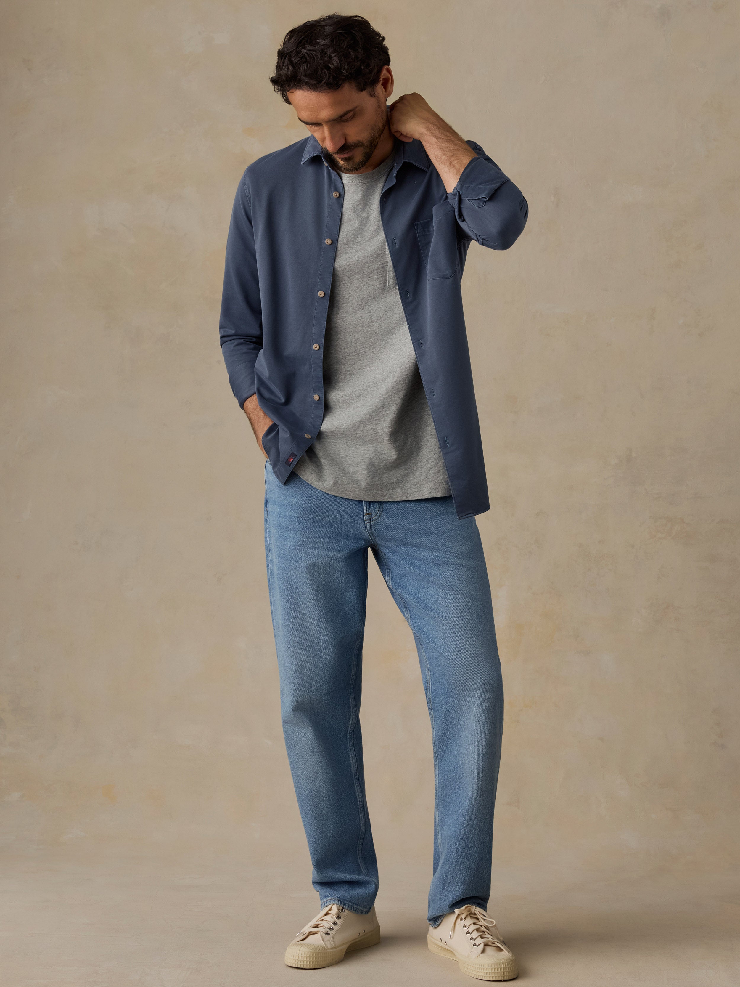 A man wearing a navy blue button-down shirt over a light gray t-shirt, paired with blue jeans and beige sneakers, standing against a textured beige studio backdrop.