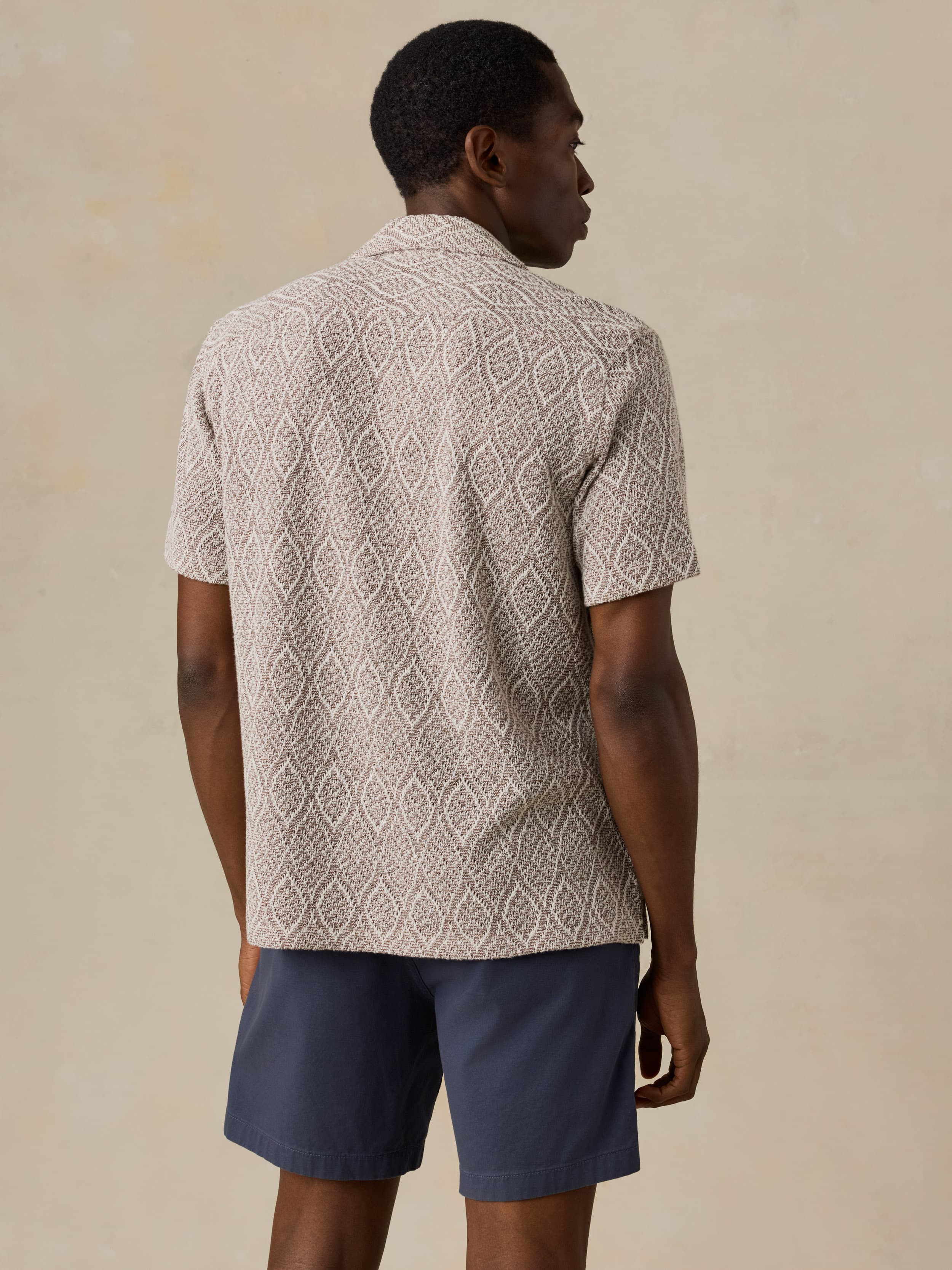 Man wearing a brown patterned shirt and blue shorts against a beige background