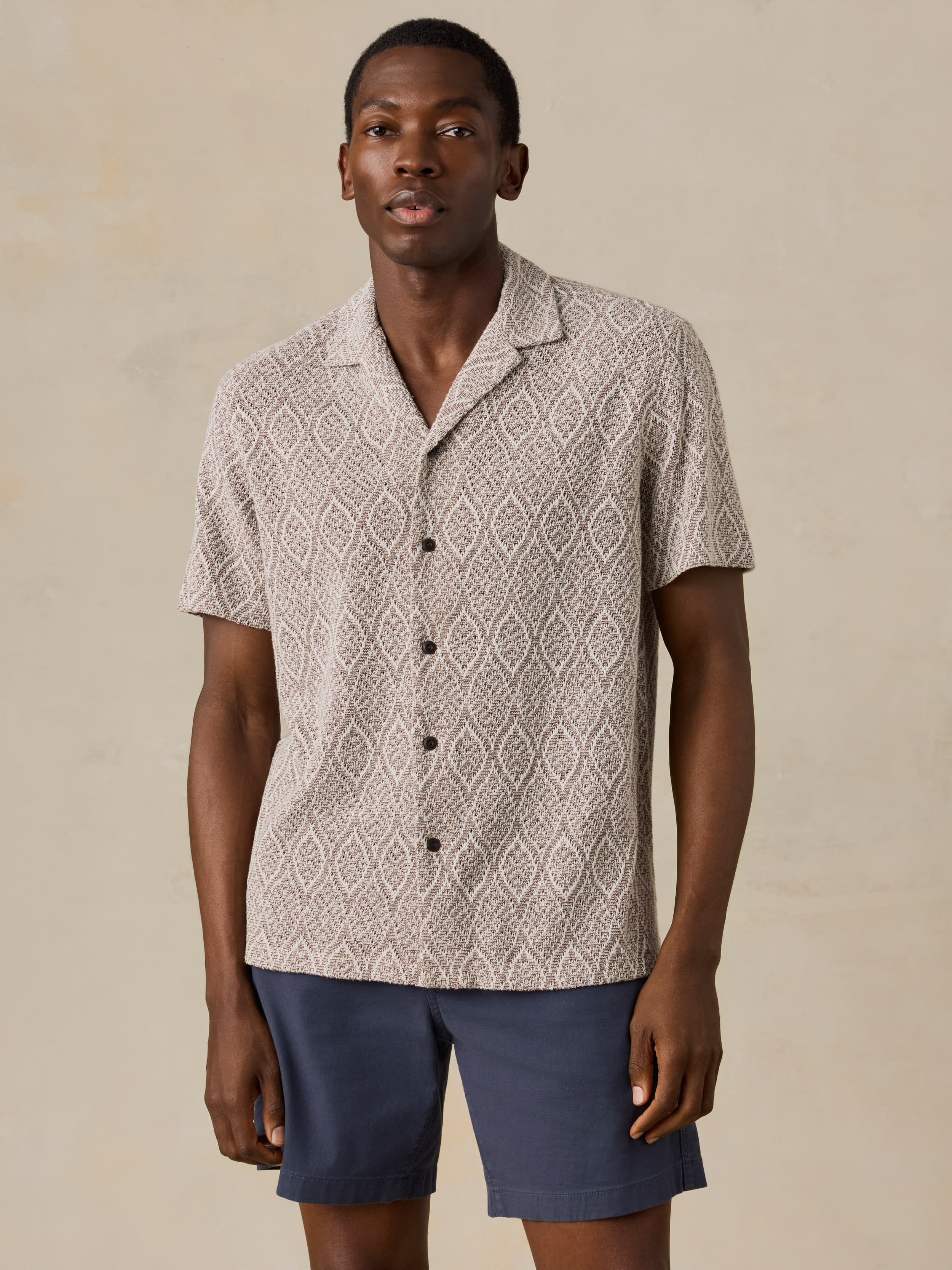 Man wearing a brown patterned shirt and blue shorts against a beige background