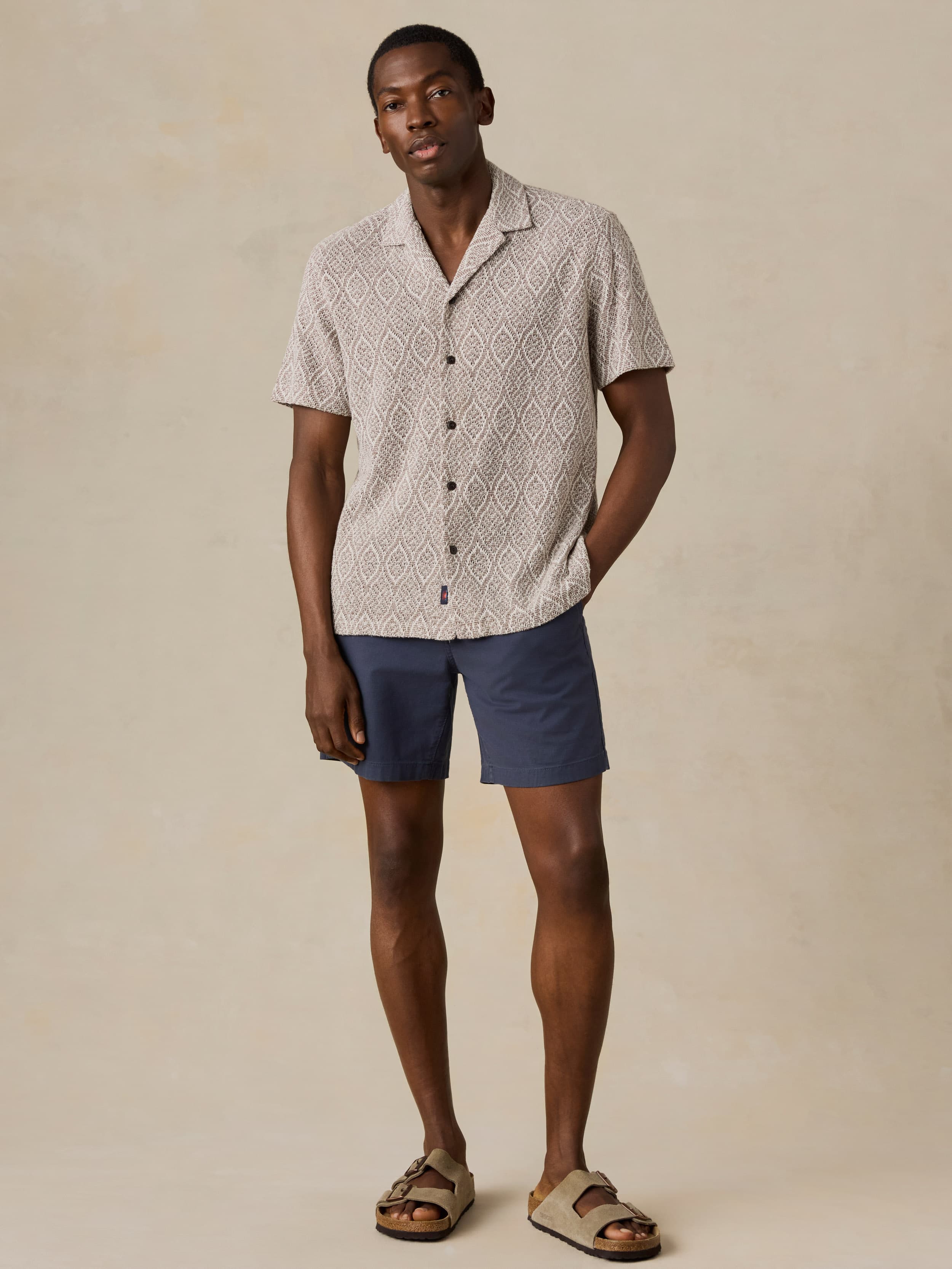 Man wearing a brown patterned shirt and blue shorts against a beige background