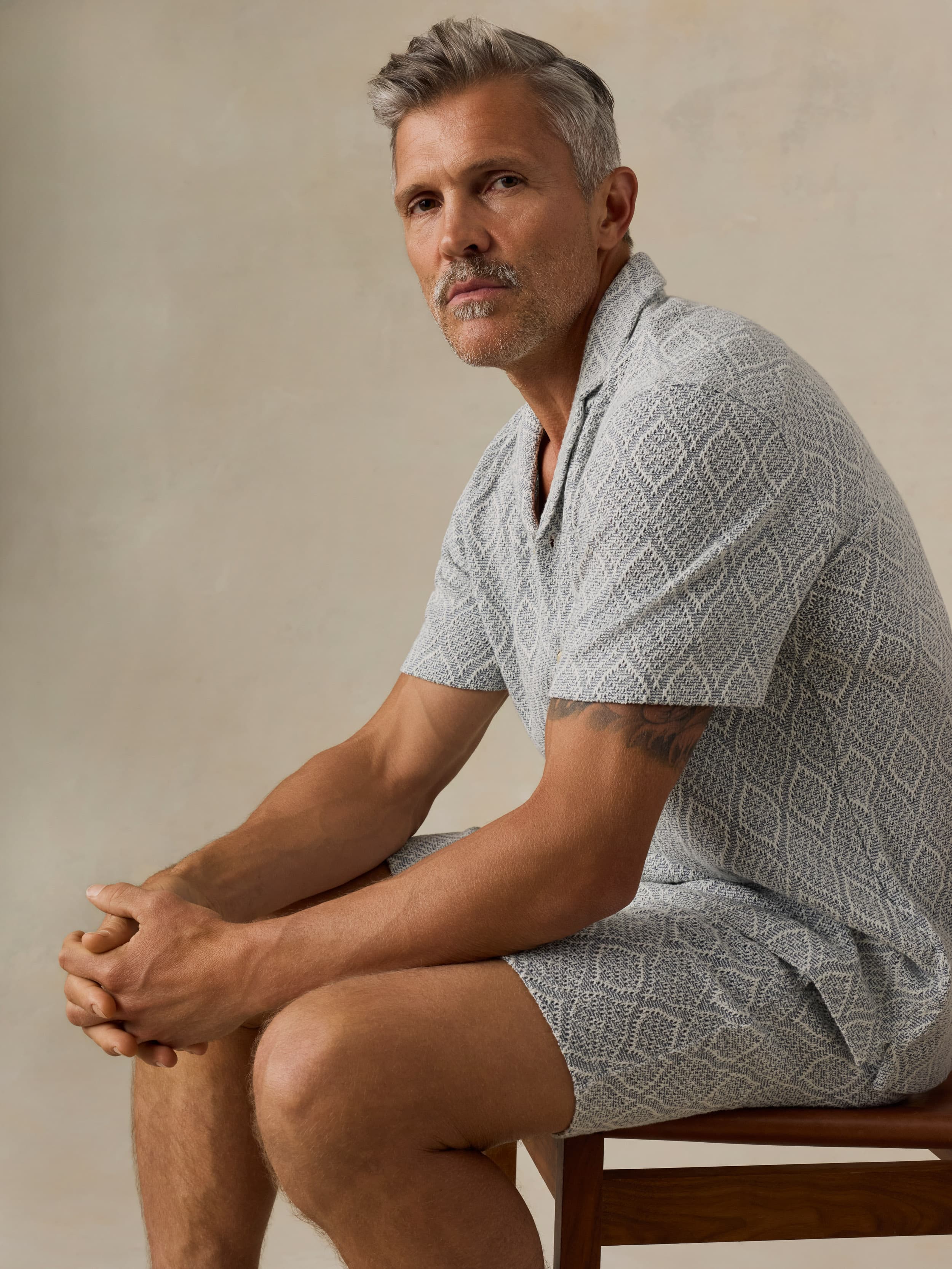 Man sitting on a wooden chair wearing a patterned light blue shirt and matching shorts against a beige background