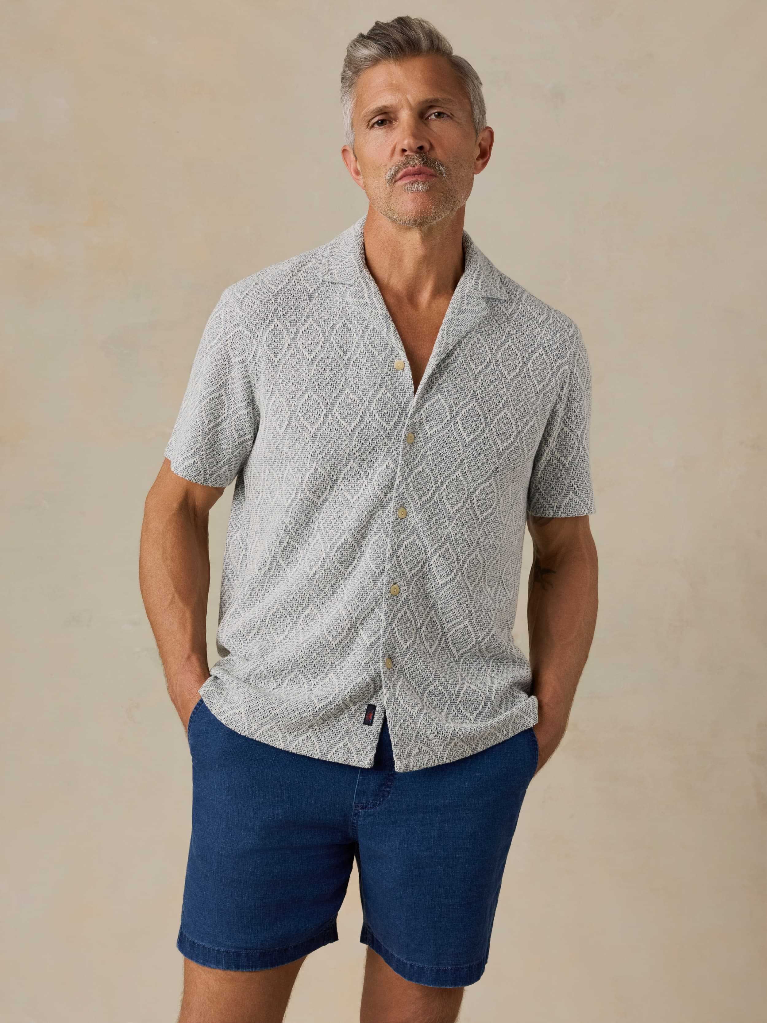 Man wearing a patterned light blue shirt and blue shorts against a beige background