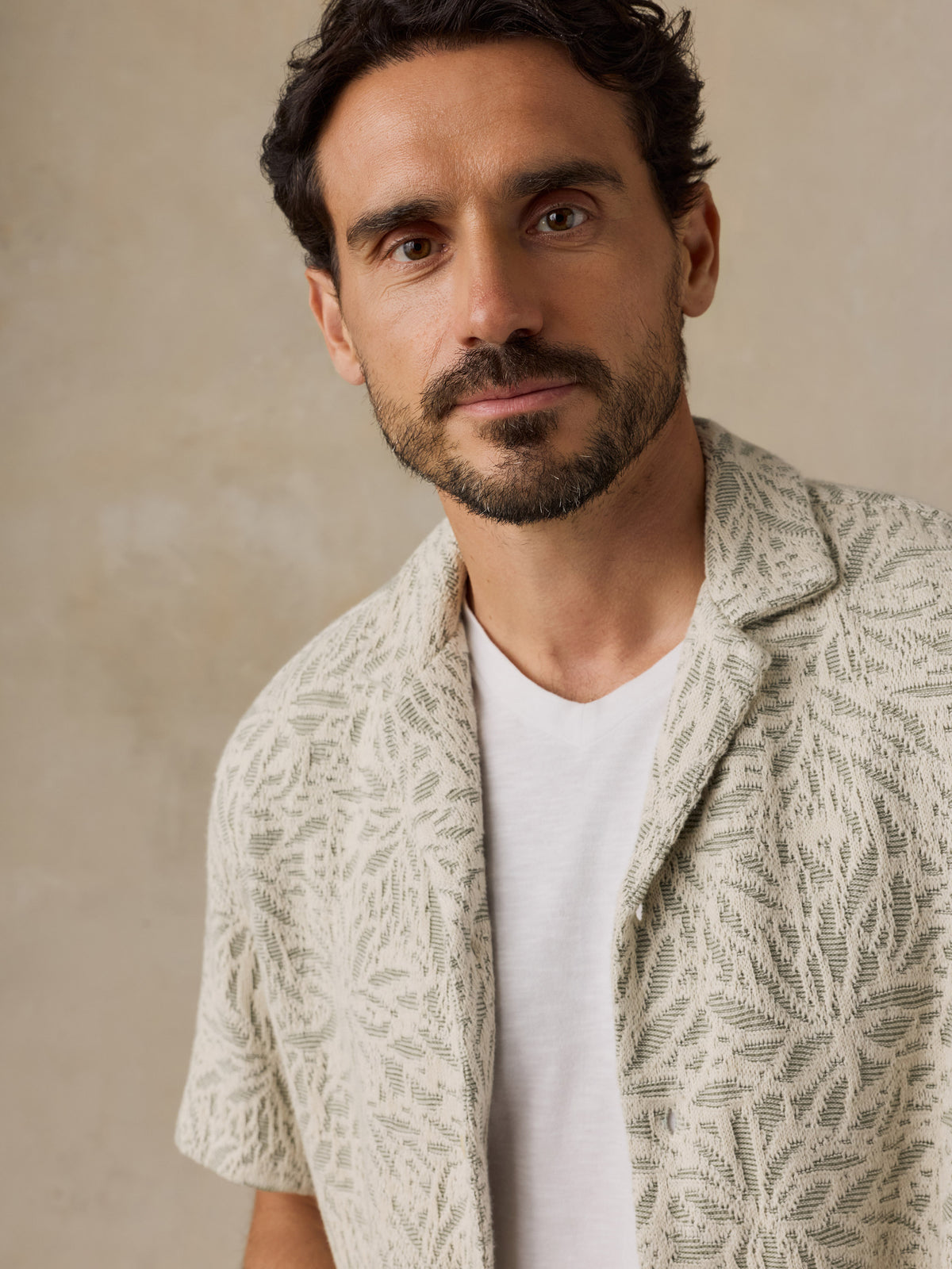 A man wearing a textured, short-sleeve cream-colored knit shirt with a leaf-like pattern over a white t-shirt, set against a neutral beige background., image 4 of 7