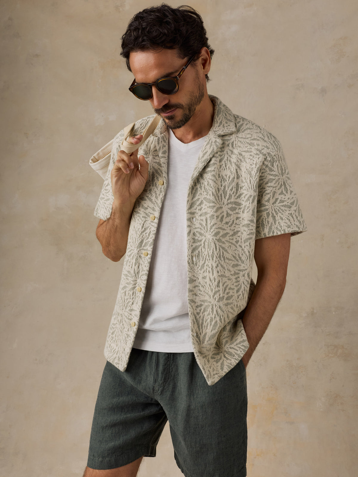 A man wearing a short-sleeve button-up shirt with a beige and white botanical leaf pattern, worn open over a white t-shirt and dark gray shorts, posing against a textured beige background., image 7 of 7