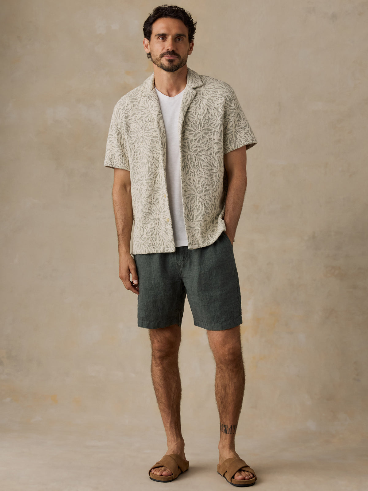 A man wearing a short-sleeve button-up shirt with a beige and brown abstract pattern, layered over a white t-shirt, dark gray shorts, and brown leather slide sandals, standing against a textured beige studio backdrop., image 6 of 7