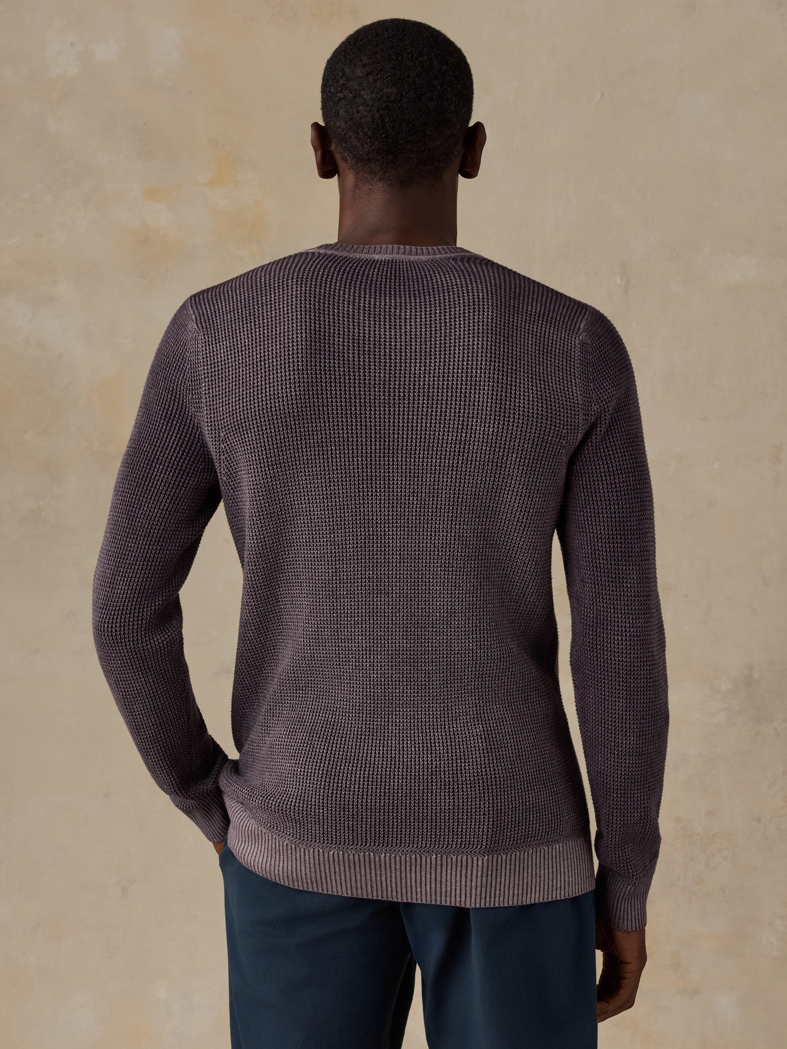 Man wearing a purple knit sweater and navy chino pants against a beige background