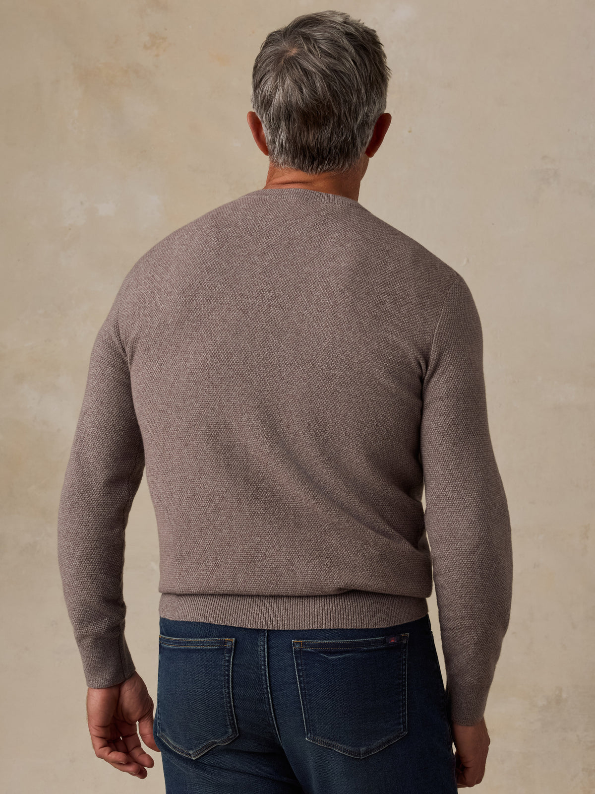 A man viewed from behind wearing a taupe brown knit crewneck sweater and dark blue jeans, standing against a textured beige wall., image 2 of 5