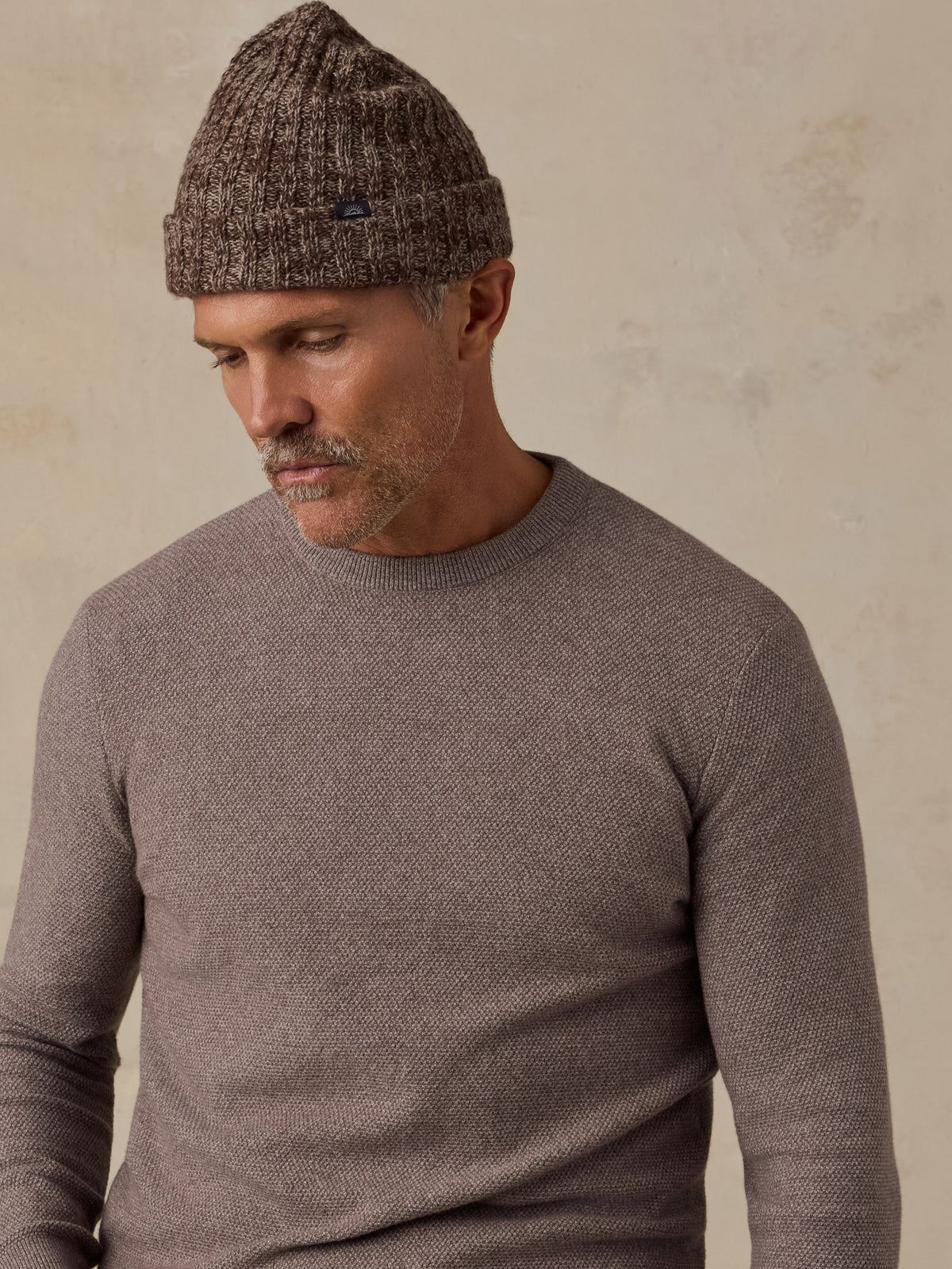 A man wearing a brown knit beanie and a taupe-gray long-sleeve crewneck sweater, photographed against a textured beige wall., image 3 of 5