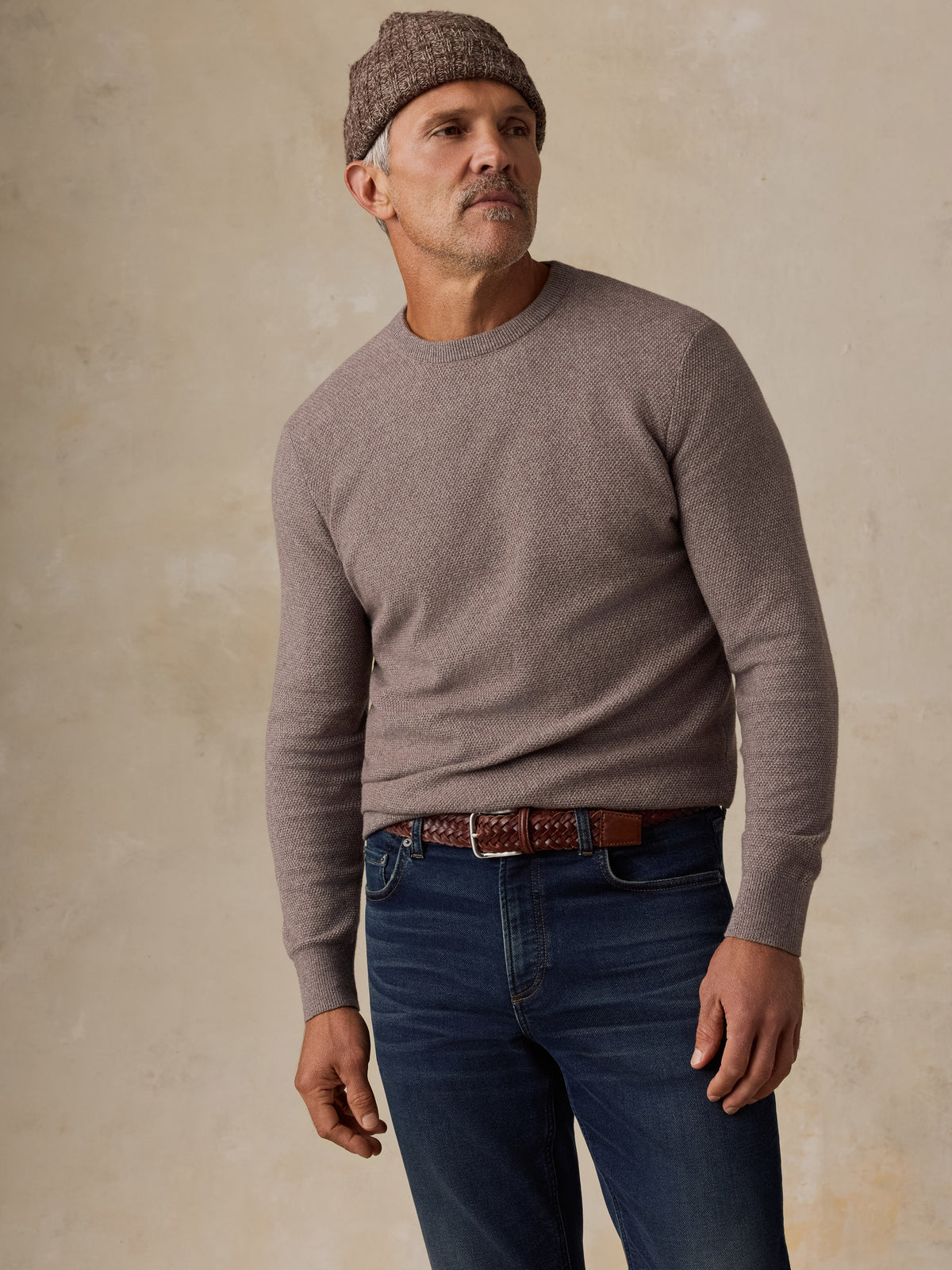 A man wearing a taupe crewneck knit sweater, blue jeans, a brown leather belt, and a brown knit beanie, standing against a textured beige studio backdrop., image 4 of 5