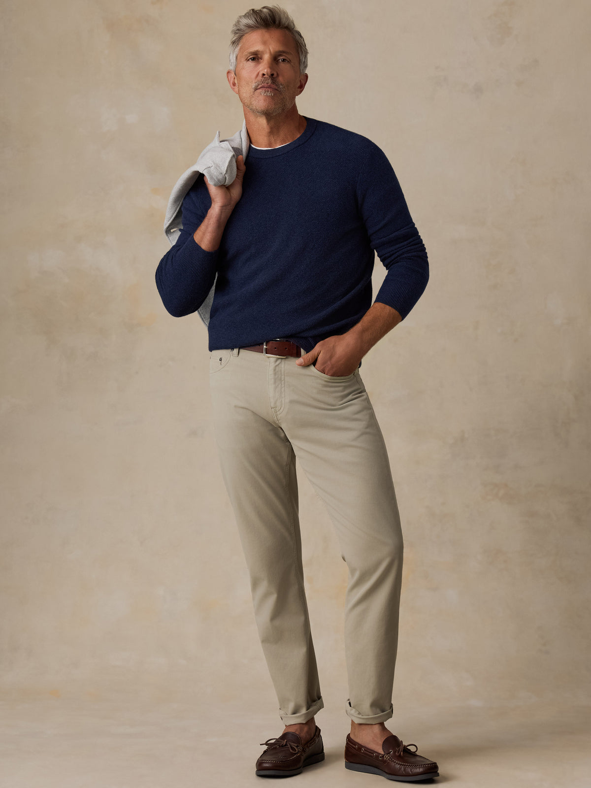 A man stands against a textured beige studio backdrop wearing a navy long-sleeve crewneck sweater, light beige trousers, and dark brown boat shoes, with a light gray shirt draped over his shoulder., image 5 of 5