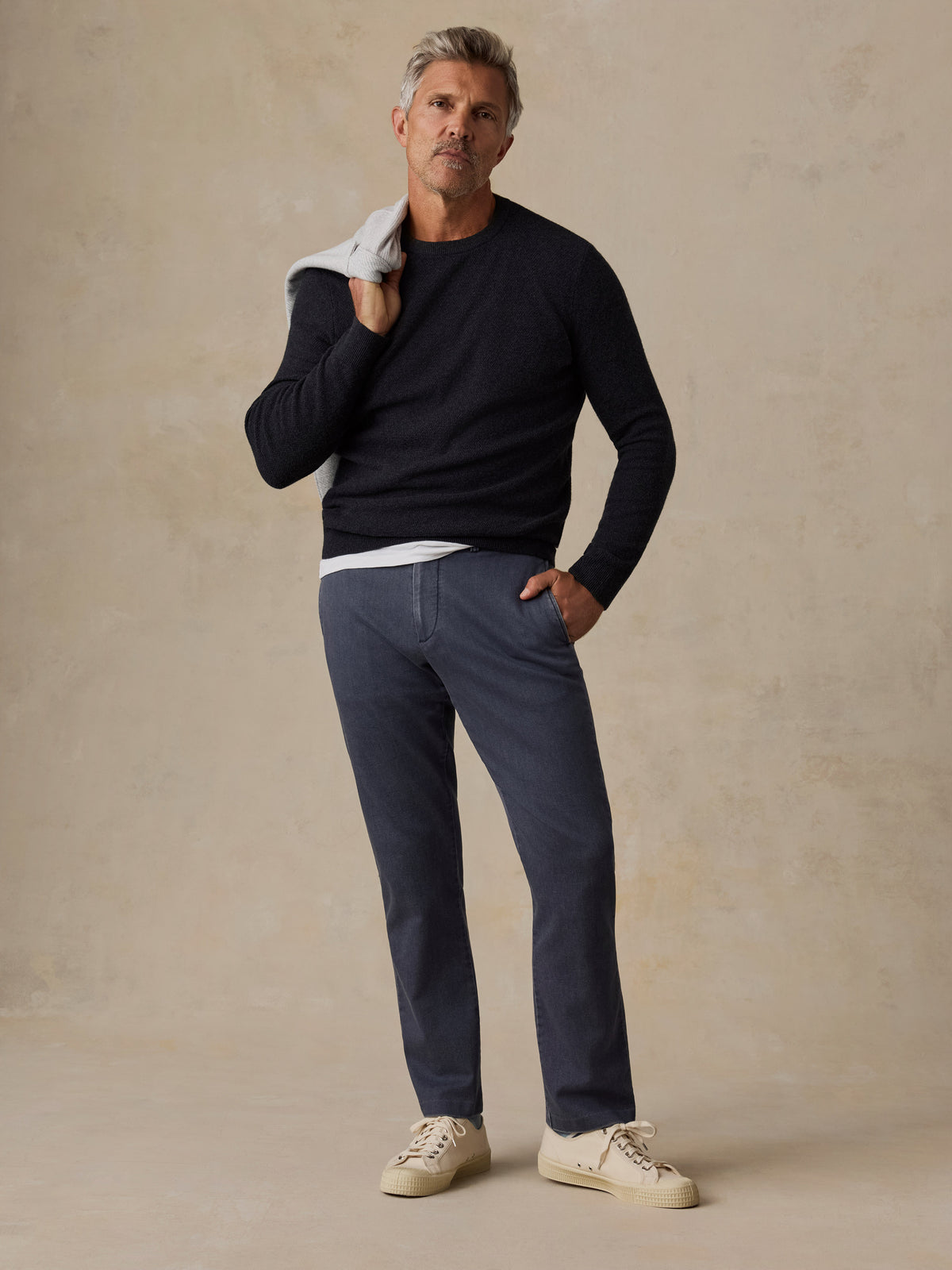 A man modeling a dark navy long-sleeve knit sweater, charcoal blue trousers, and cream sneakers, image 2 of 7