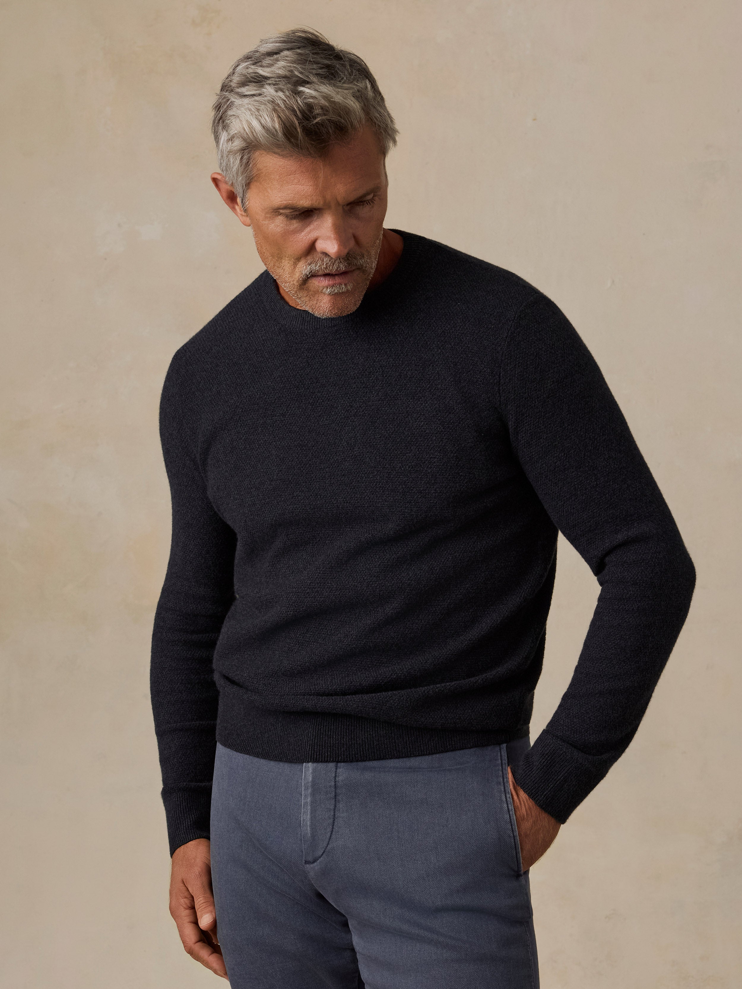 A man wearing a dark navy blue knit crewneck sweater and slate blue trousers