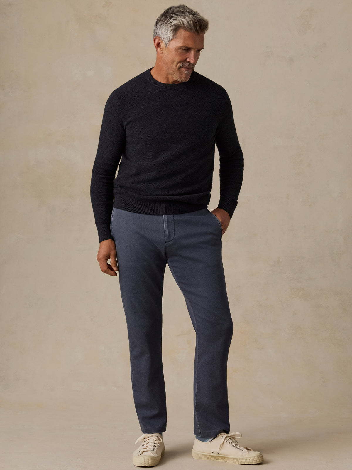 A man wearing a dark navy crewneck sweater, slate blue jeans, and off-white sneakers, image 5 of 7