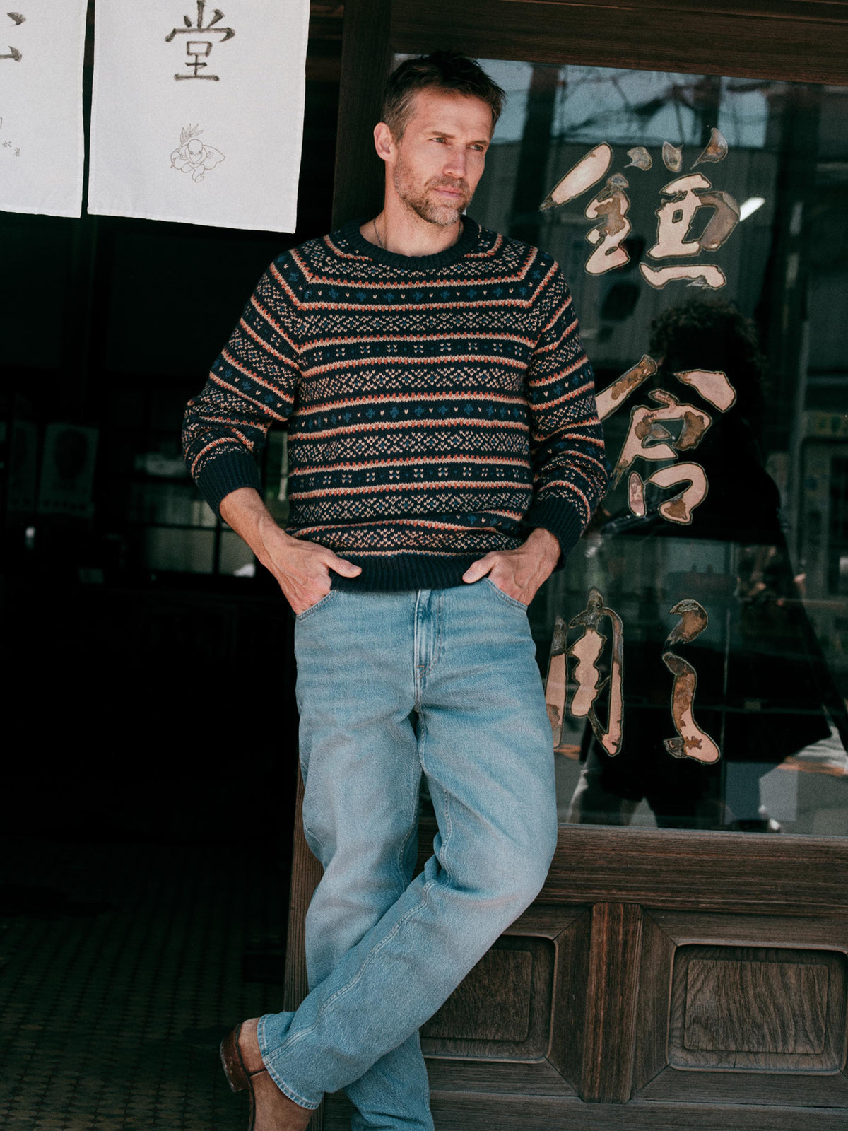 Man wearing a knit fairisle sweater and blue jeans standing outside of a Japanese store, image 1 of 6