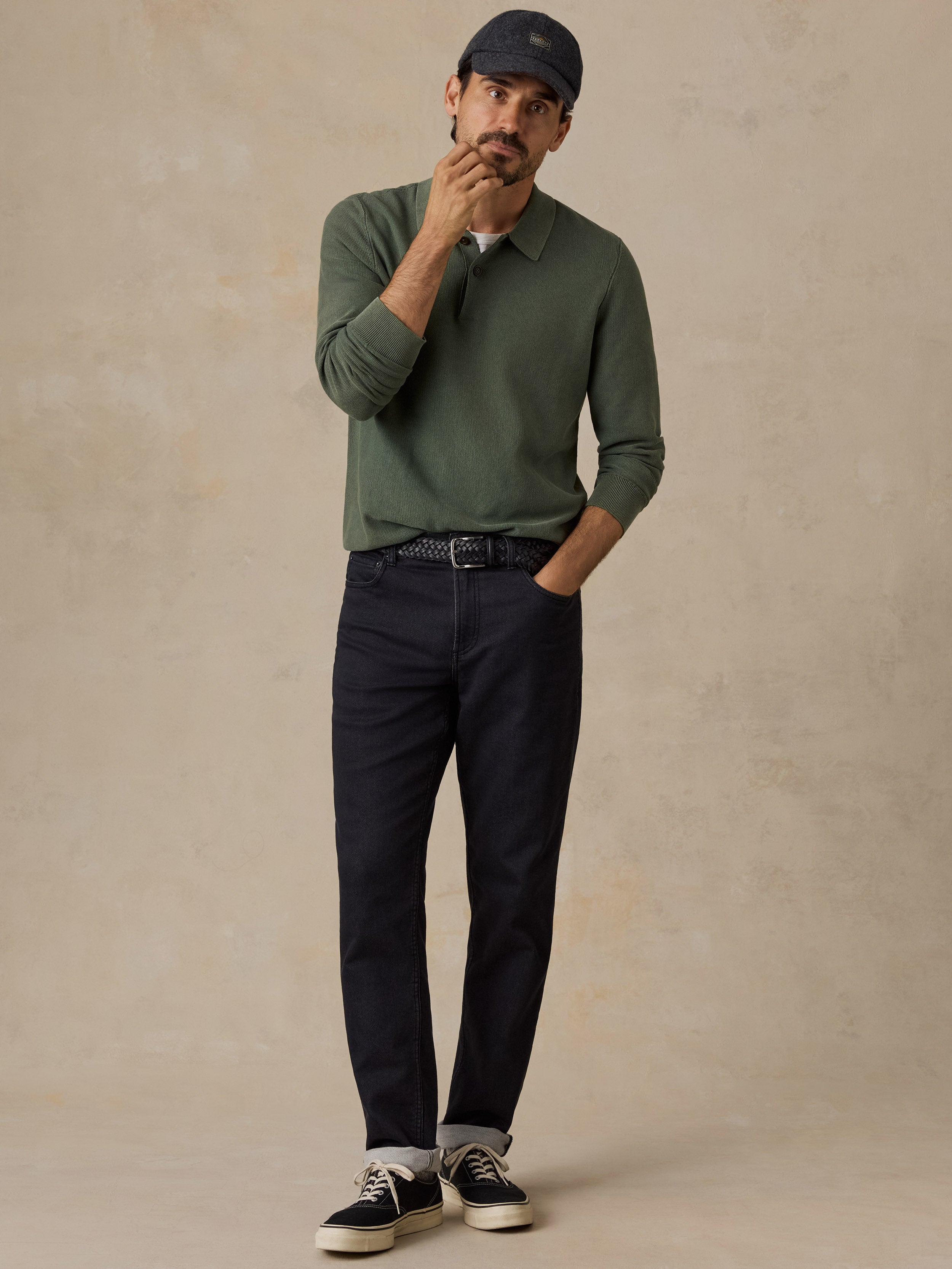 A man wearing an olive green long-sleeve polo shirt, dark navy pants, a navy baseball cap, and black sneakers with white soles, standing against a textured beige studio backdrop.