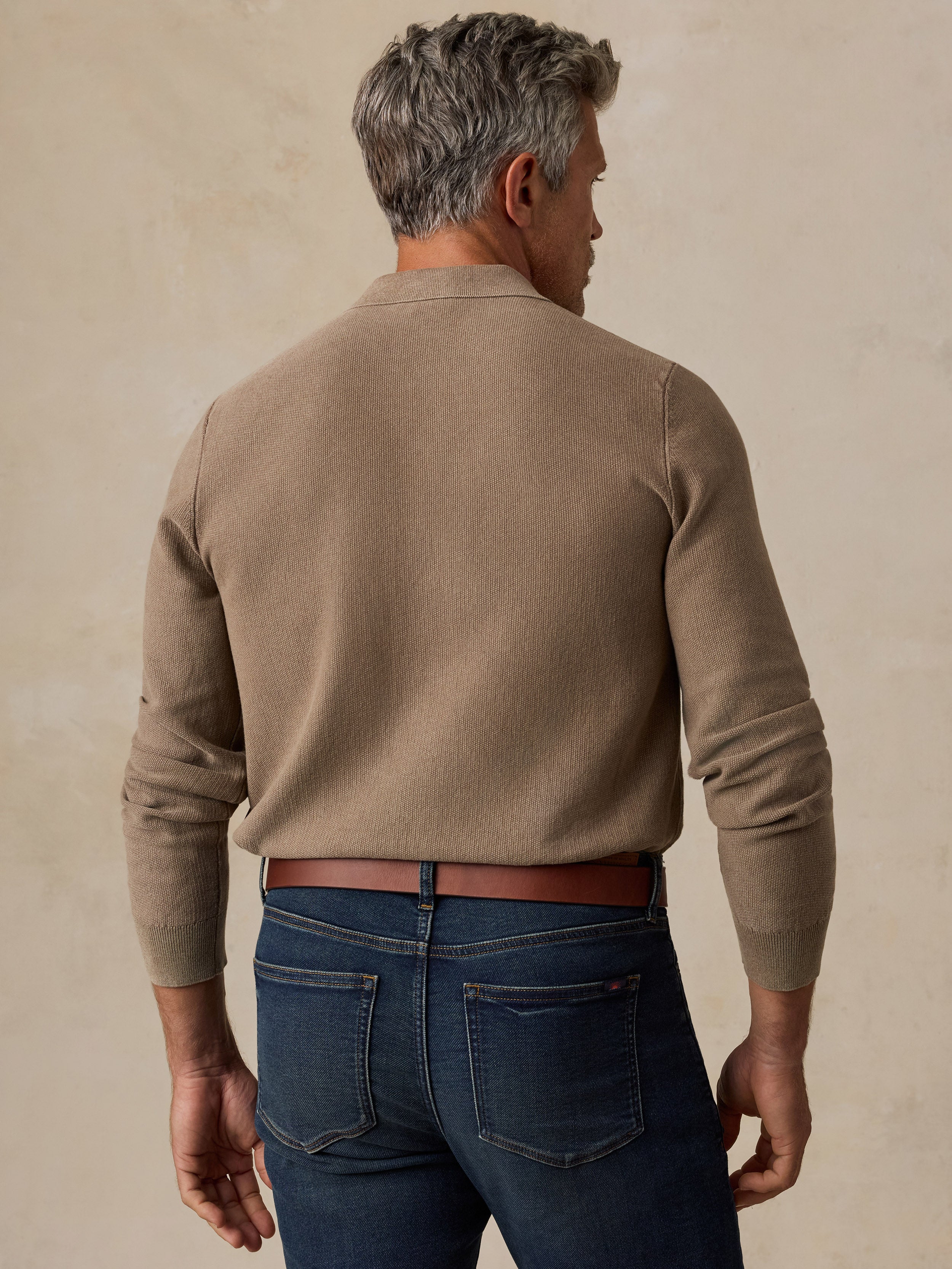 A man viewed from behind wearing a taupe brown knit sweater and dark blue jeans, standing against a textured beige background.