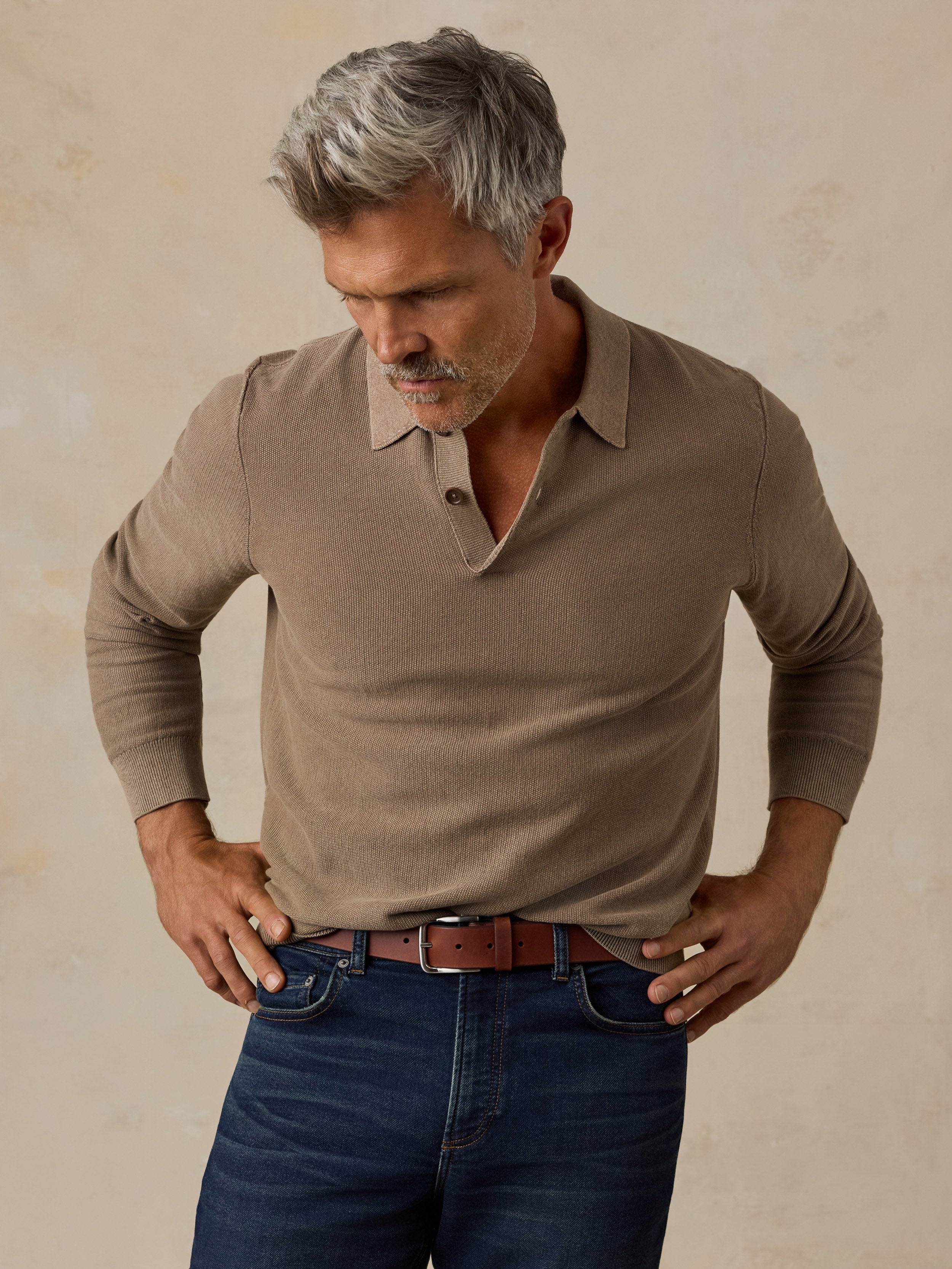 A man with gray hair wearing a taupe brown long-sleeve knit polo shirt and dark blue jeans, standing against a textured beige background.