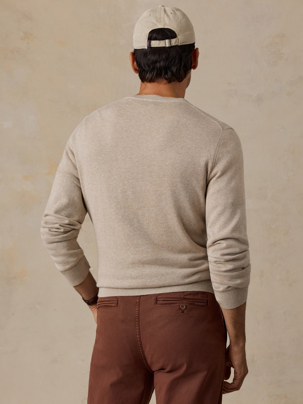 A man wearing a light beige knit crewneck sweater, brown pants, and a beige baseball cap.