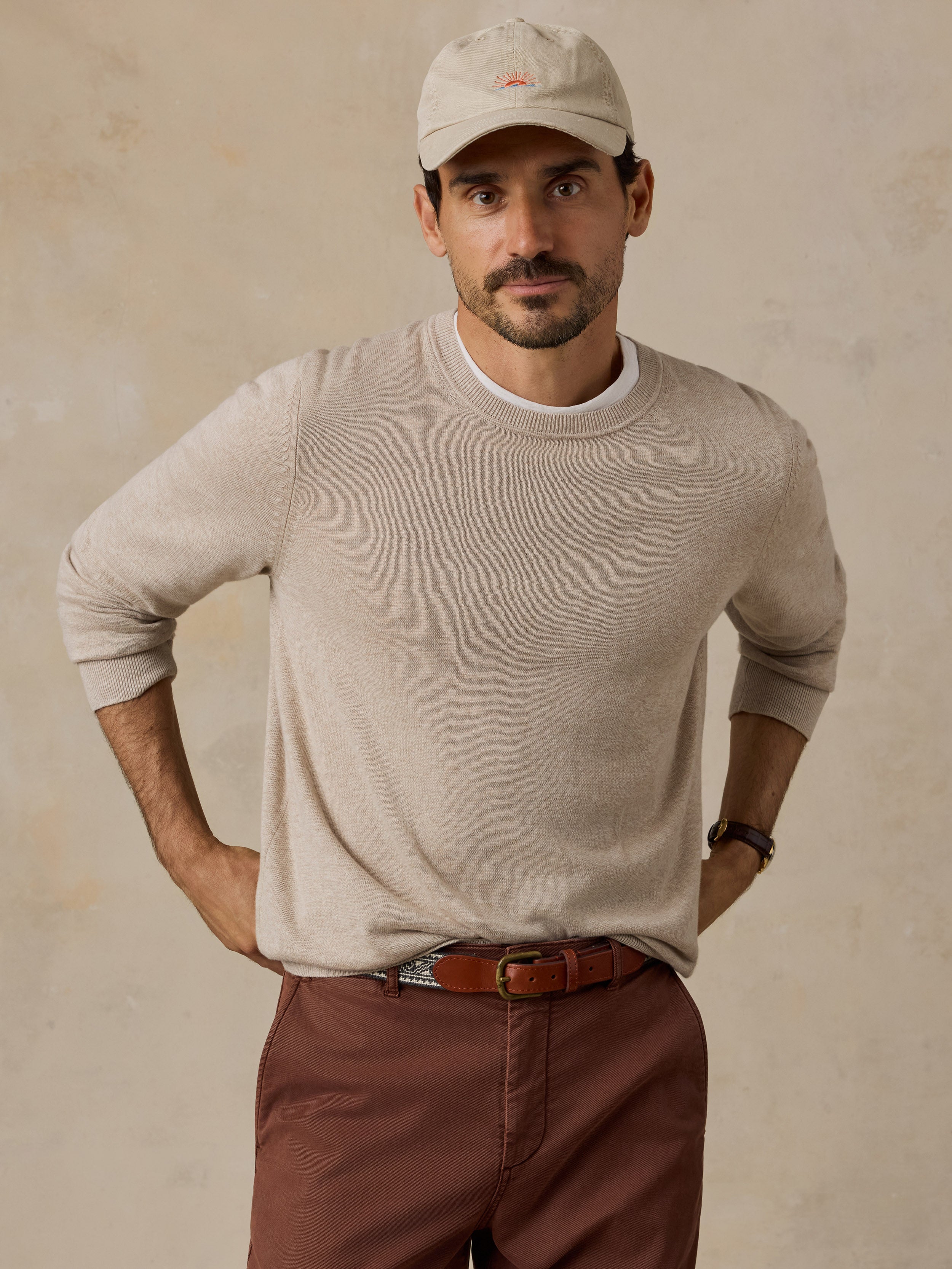 A man wearing a light beige crewneck sweater, a matching beige baseball cap, brown pants, and a brown belt.
