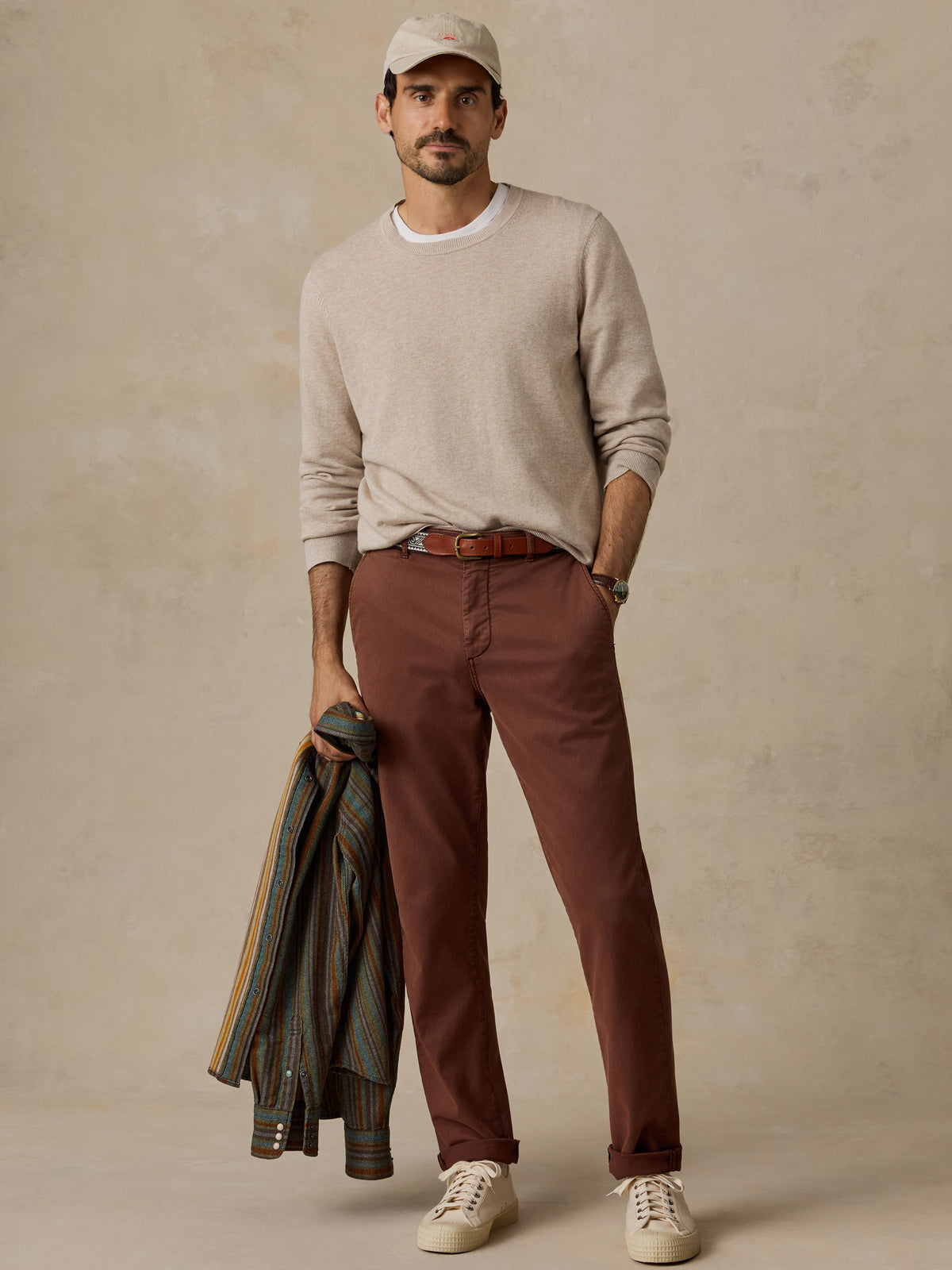 A man wearing a light beige crewneck sweater, rust-brown pants, a tan baseball cap, white sneakers.